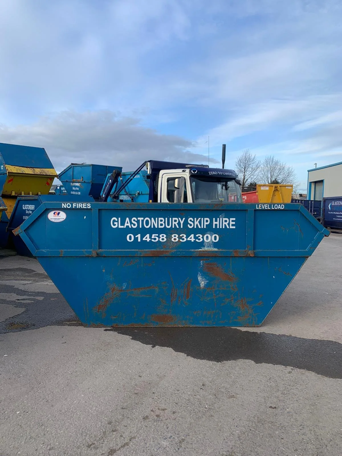Blue skip hire container with white lettering reading 'Glastonbury Skip Hire' and contact number, parked outdoors with other containers and a small truck, under a cloudy sky.