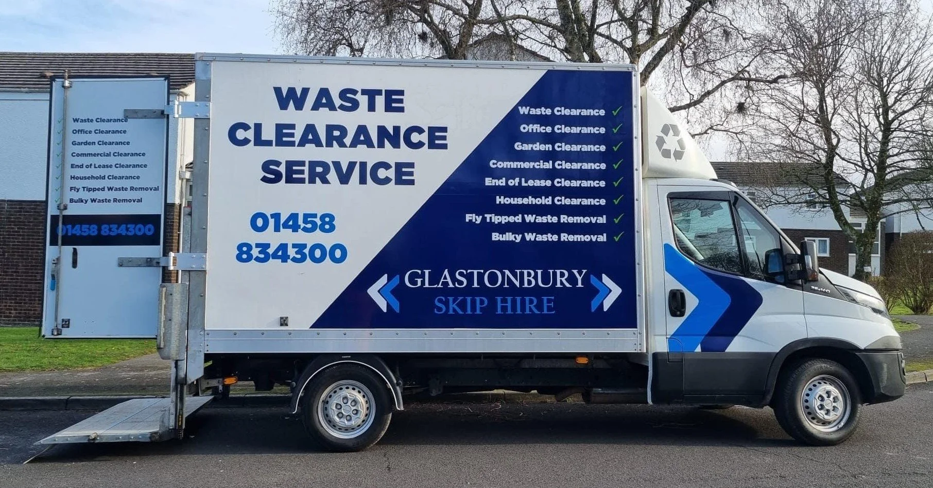 A white waste clearance service truck with blue graphics and text on the side and back, parked on a residential street. The truck features services such as waste clearance, office clearance, garden clearance, commercial clearance, end of lease clearance, household clearance, fly tipped waste removal, and bulky waste removal. The contact number is 01458 834300. The logo indicates it is operated by Glastonbury Skip Hire.