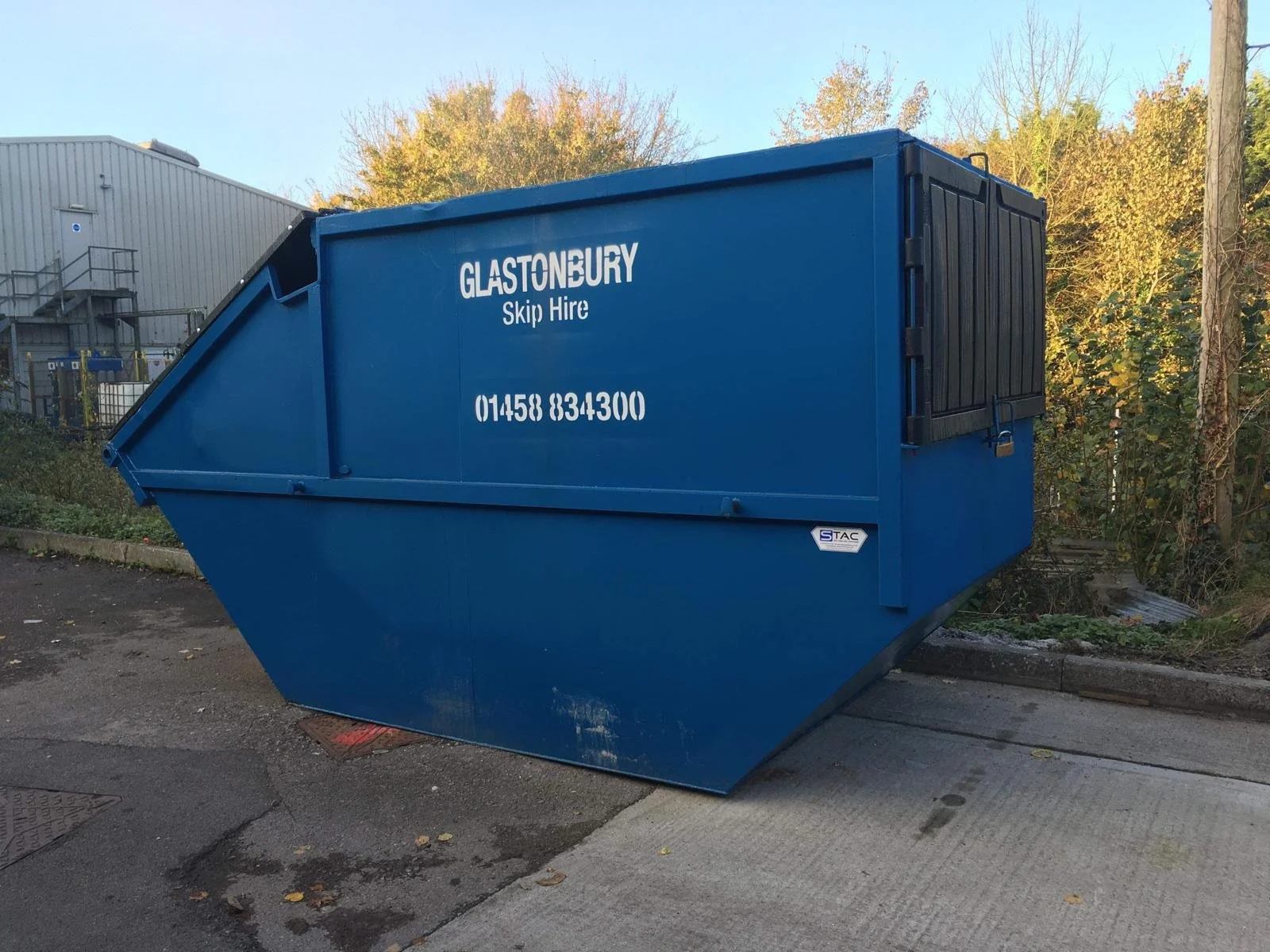 Blue skip hire waste container with contact phone number and company name, situated outdoors on a street near some bushes and a building.