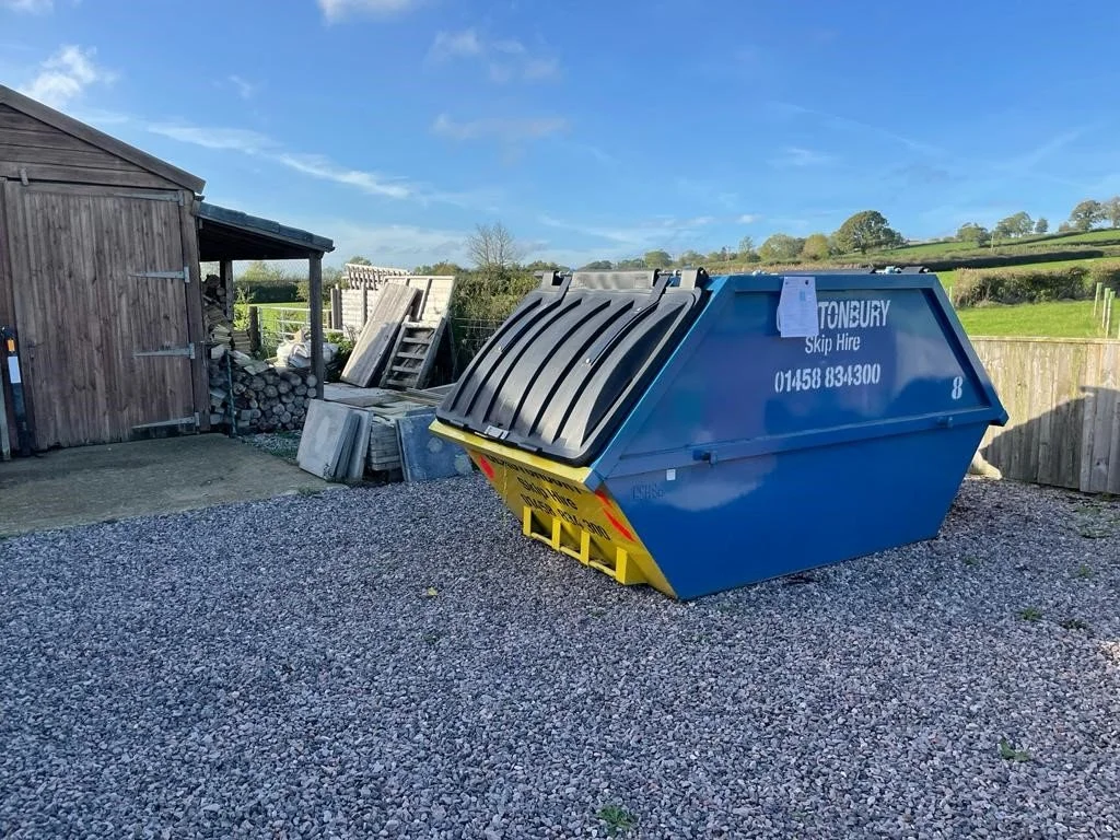 A blue skip container labeled 'Tonbury Skip Hire' on a gravel area outside a shed and wooden fencing, with trees and fields in the background.