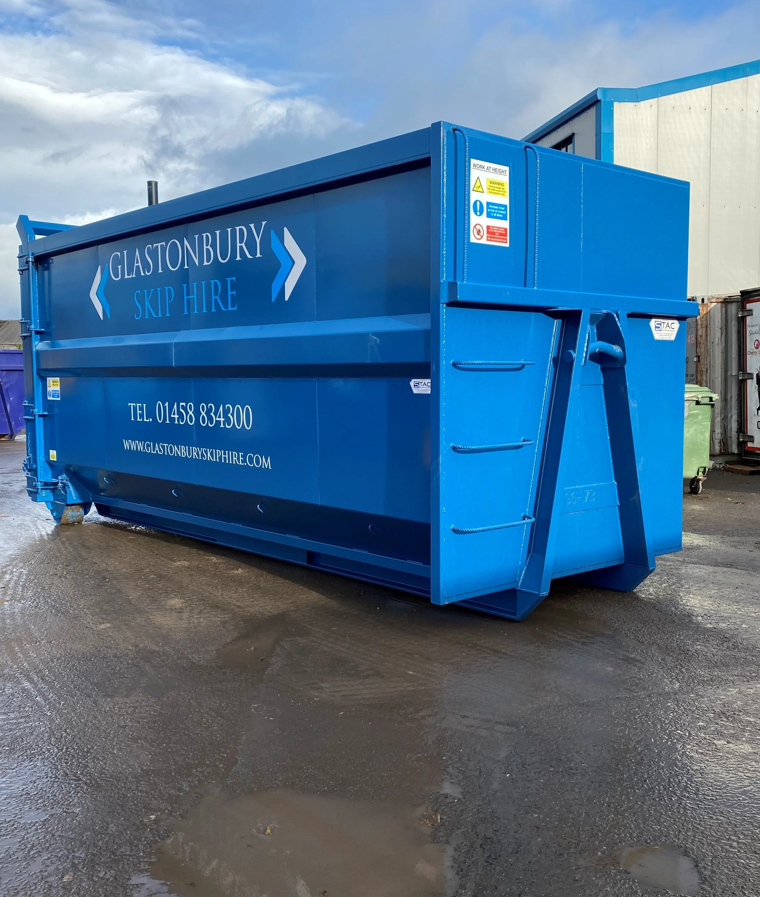 Blue skip hire container with company information and safety signage, located outdoors on wet ground, with clouds in the sky and industrial buildings in the background.