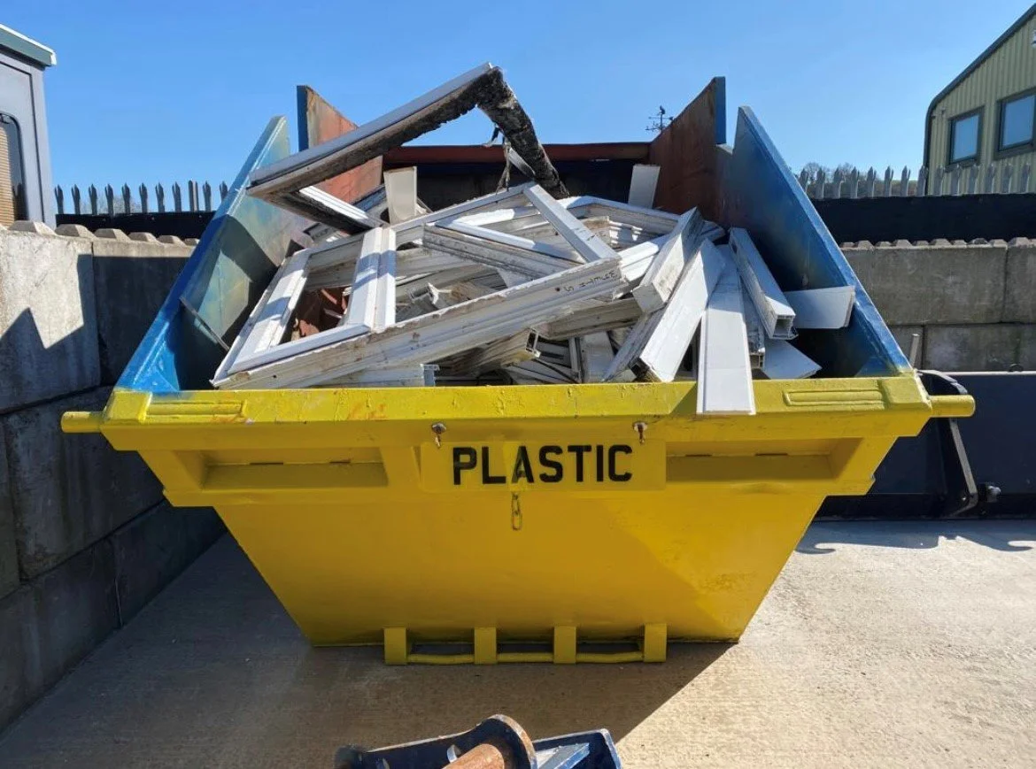 A large yellow dumpster labeled 'PLASTIC' filled with various discarded white window frames and glass panes outside on a concrete surface.