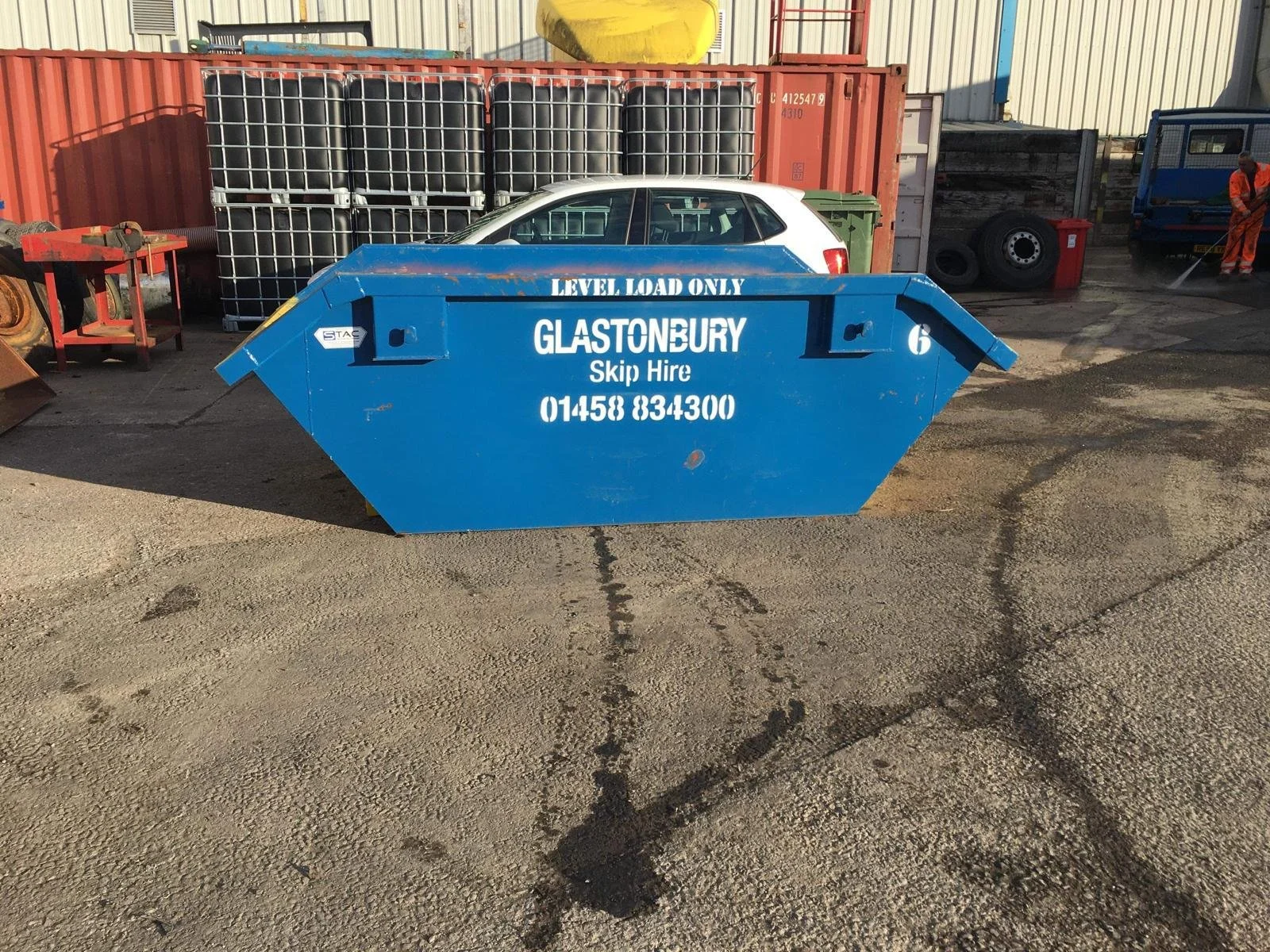 Blue skip bin with white text advertising Glastonbury Skip Hire, a phone number, and the words 'Level Load Only', placed on a concrete ground outside a storage yard with containers, barrels, and a person disinfecting in the background.