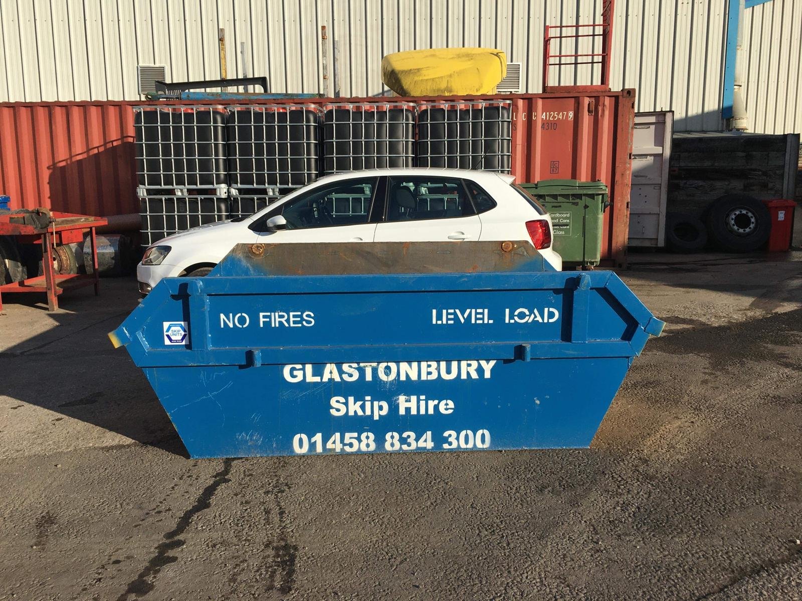 Blue skip bin with white text reading 'NO FIRES LEVEL LOAD GLASTONBURY Skip Hire 01458 834 300', positioned on pavement with a white car behind it and industrial containers in the background.