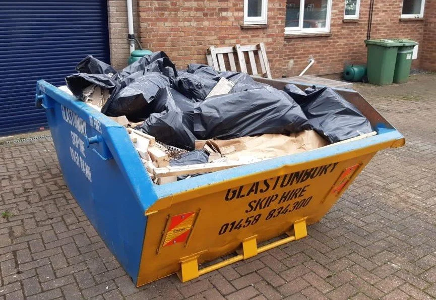 A large blue and yellow dumpster filled with black garbage bags and cardboard, placed on a brick-paved area outside a building with brick walls and a green trash bin nearby.