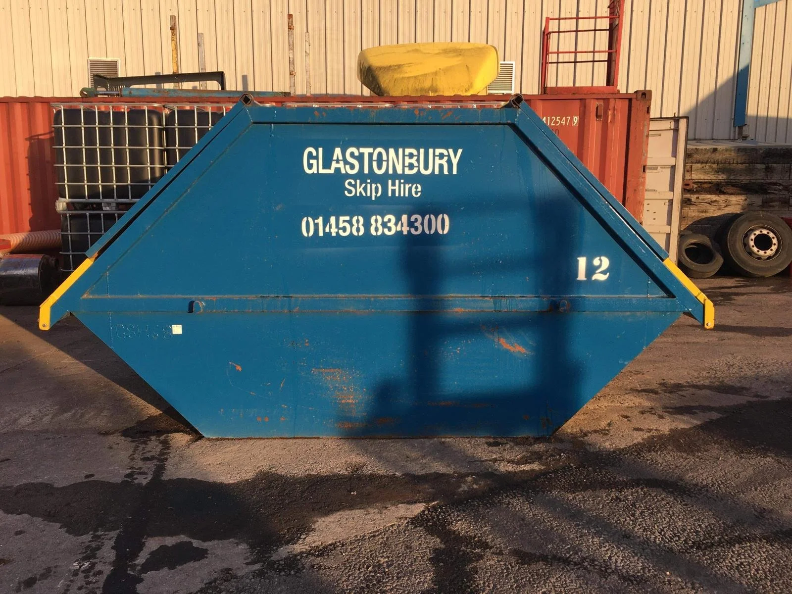Blue skip container with white text, in an industrial yard, with tires and metal racks in the background.