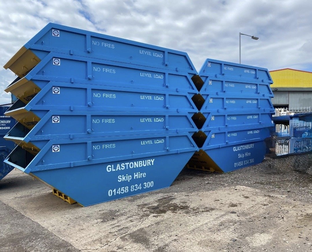Stacked blue skip bins labeled "NO FIRES" and "LEVEL LOAD" at Glastonbury Skip Hire, with a contact phone number, in an outdoor storage area.