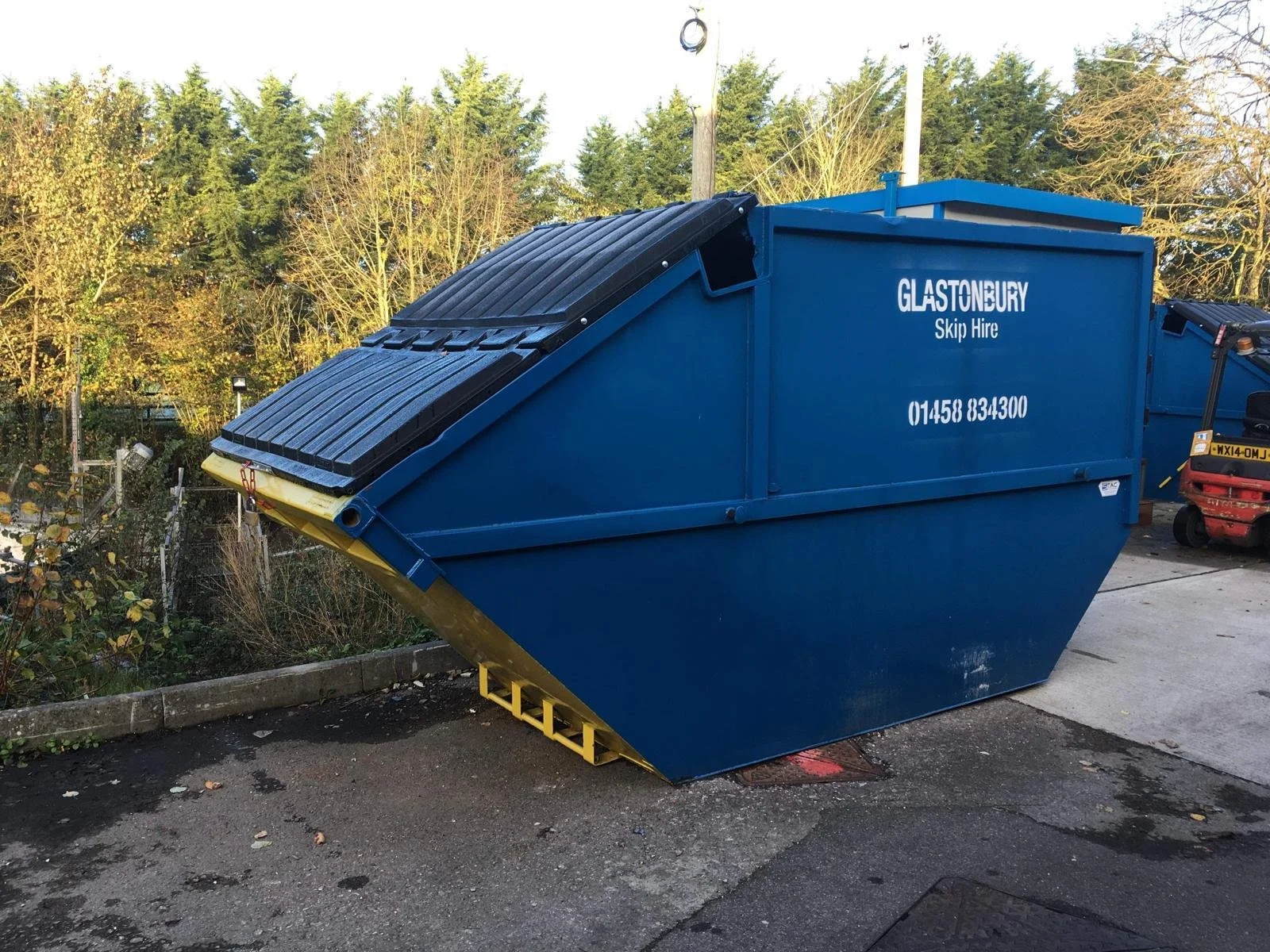 A blue skip hire waste container with a closed black lid, located outdoors on a concrete surface, with trees and a utility pole in the background.