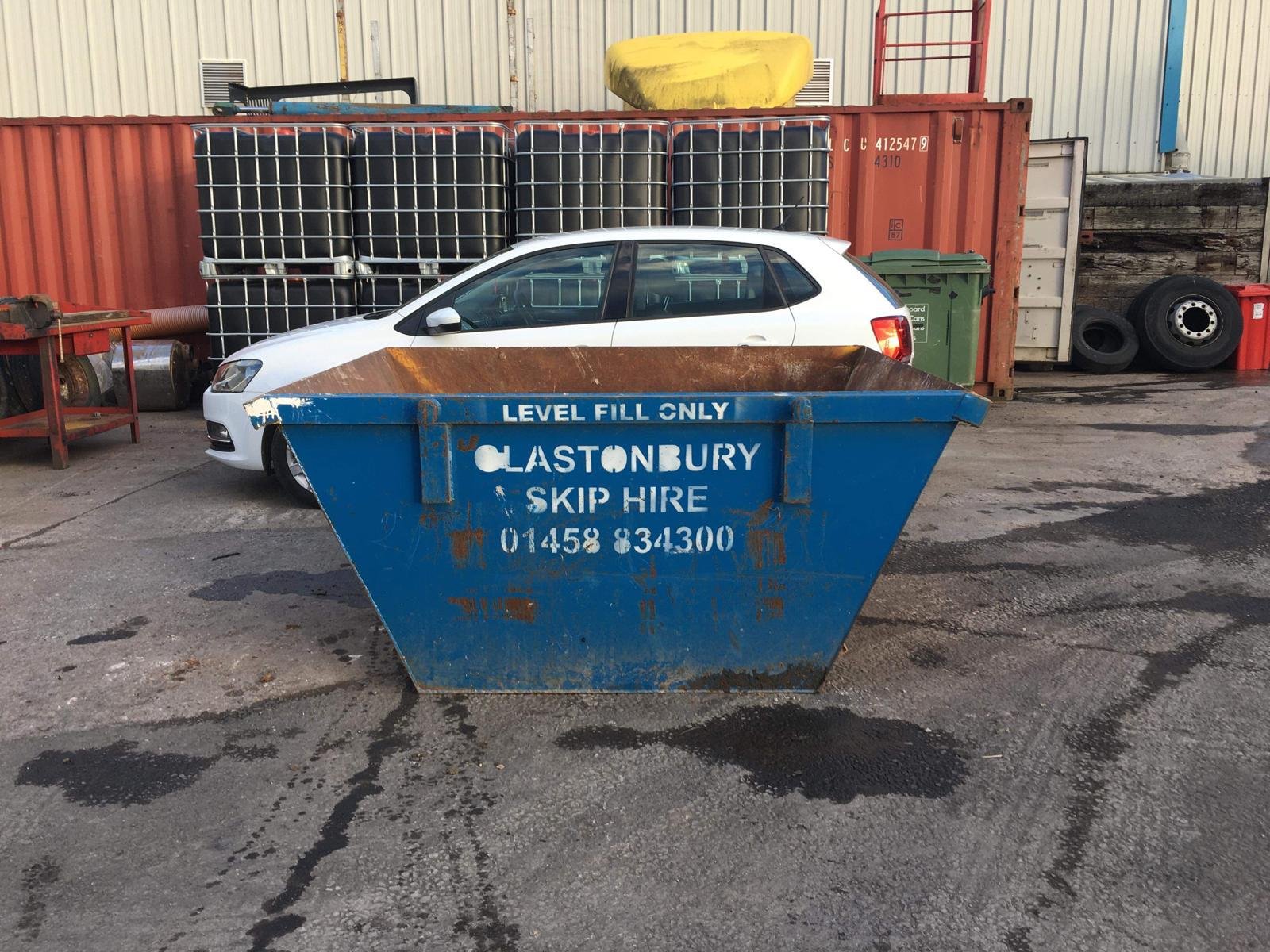 A blue skip bin with contact information for Glastonbury Skip Hire, placed in front of a white car parked in an industrial yard with containers, tires, and storage items in the background.