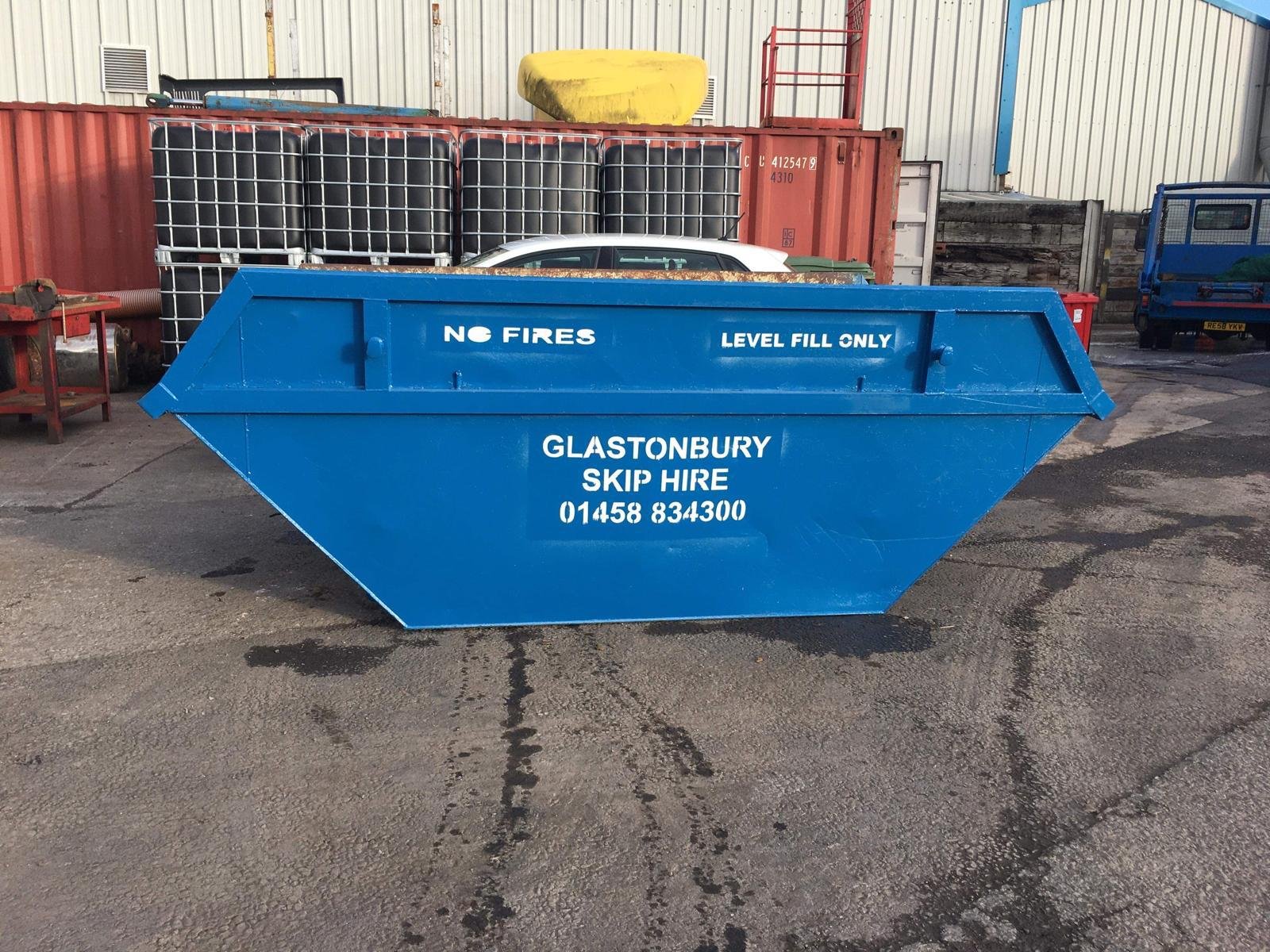 A large blue skip with white text advertising Glastonbury skip hire, placed on an asphalt surface outside a warehouse. The skip has signage stating 'NO FIRES,' 'LEVEL FILL ONLY,' and contact details.
