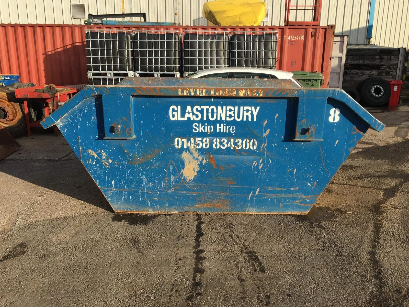 Large blue industrial skip bin with the words 'Glastonbury Skip Hire' and a phone number printed on the side, parked on a gravel surface next to other waste containers and industrial equipment.