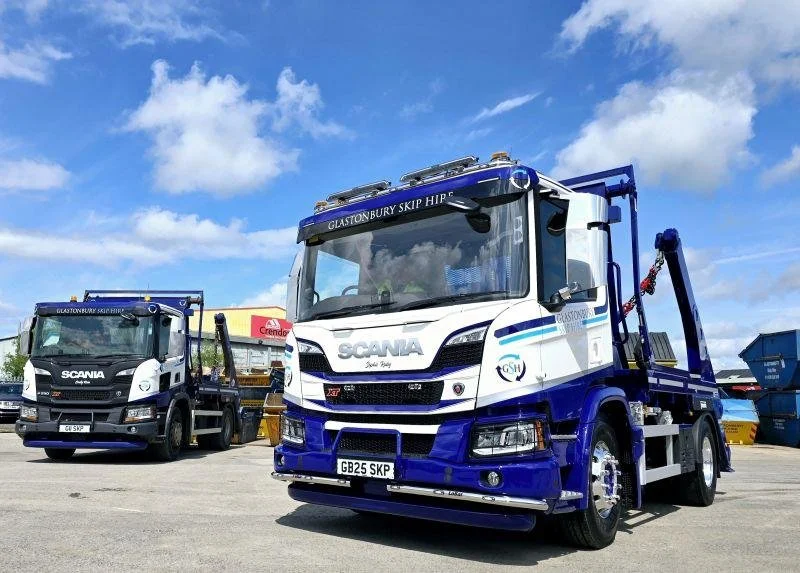Two skip loader trucks parked outdoors on a paved lot, one is white and blue with 'Glastonbury Skip Hire' written on it, under a partly cloudy sky.