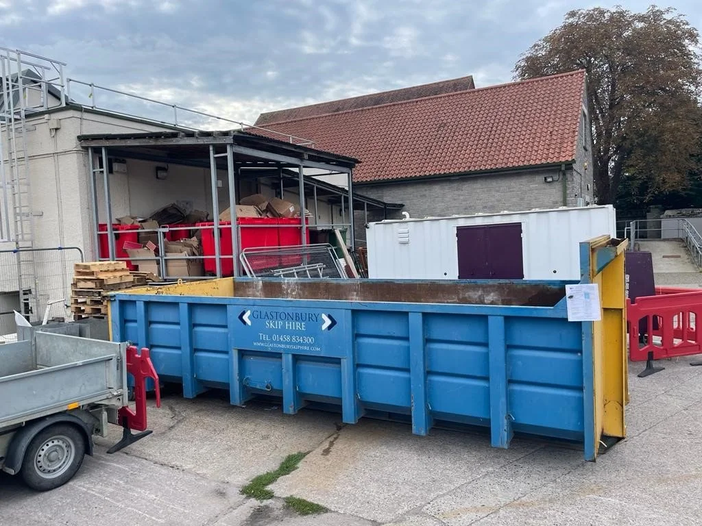 A large blue skip bin labeled 'Glastonbury Skip Hire' filled with debris, positioned outside a building with stacked wooden pallets and other waste materials nearby.