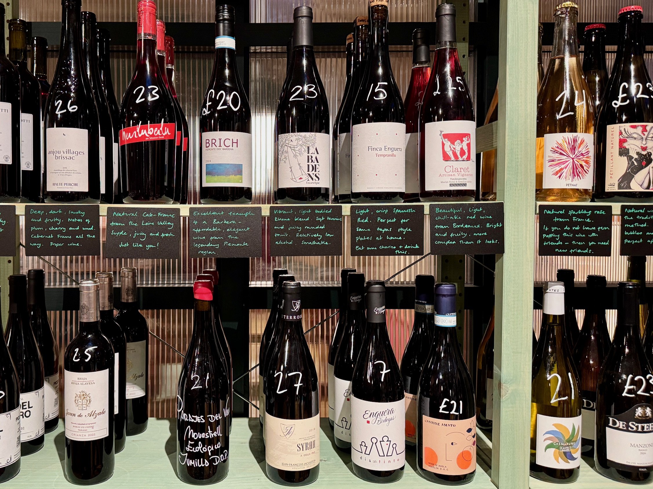 Display of various wine bottles on shelves with handwritten descriptions and prices underneath.