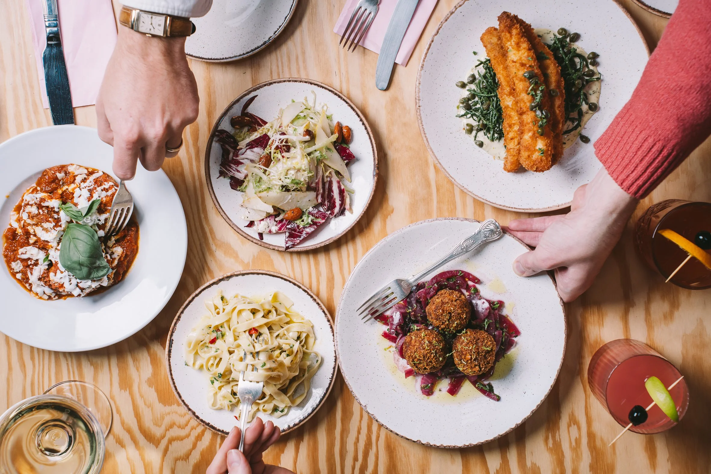 A table with various plates of food, including pasta, salad, breaded fried fish with greens, and meatballs, along with drinks and cutlery, viewed from above.