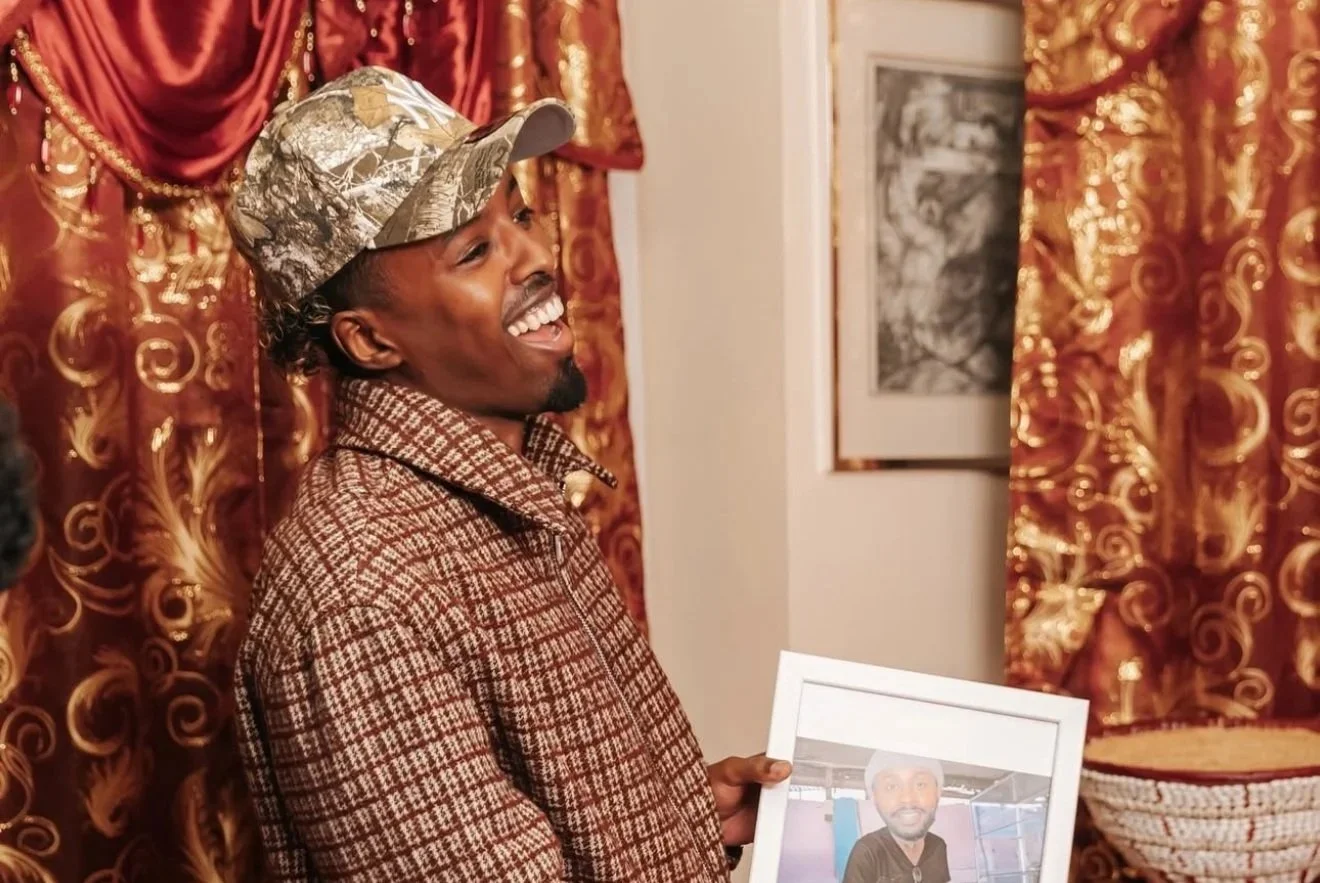 A smiling man wearing a camouflage cap and a brown checkered jacket is holding a framed photo of another man. The background features heavily decorated red and gold curtains and a framed picture on the wall.