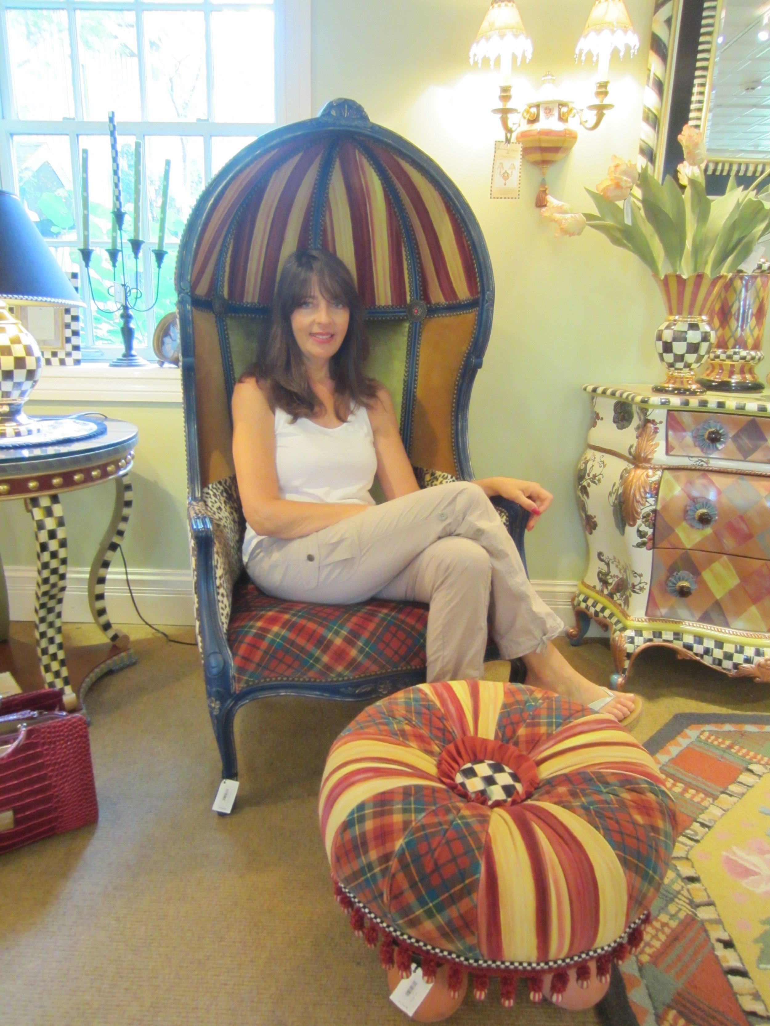 A woman seated on a colorful, high-back, striped and patterned armchair inside a decorated room with various patterned furniture and lamps.