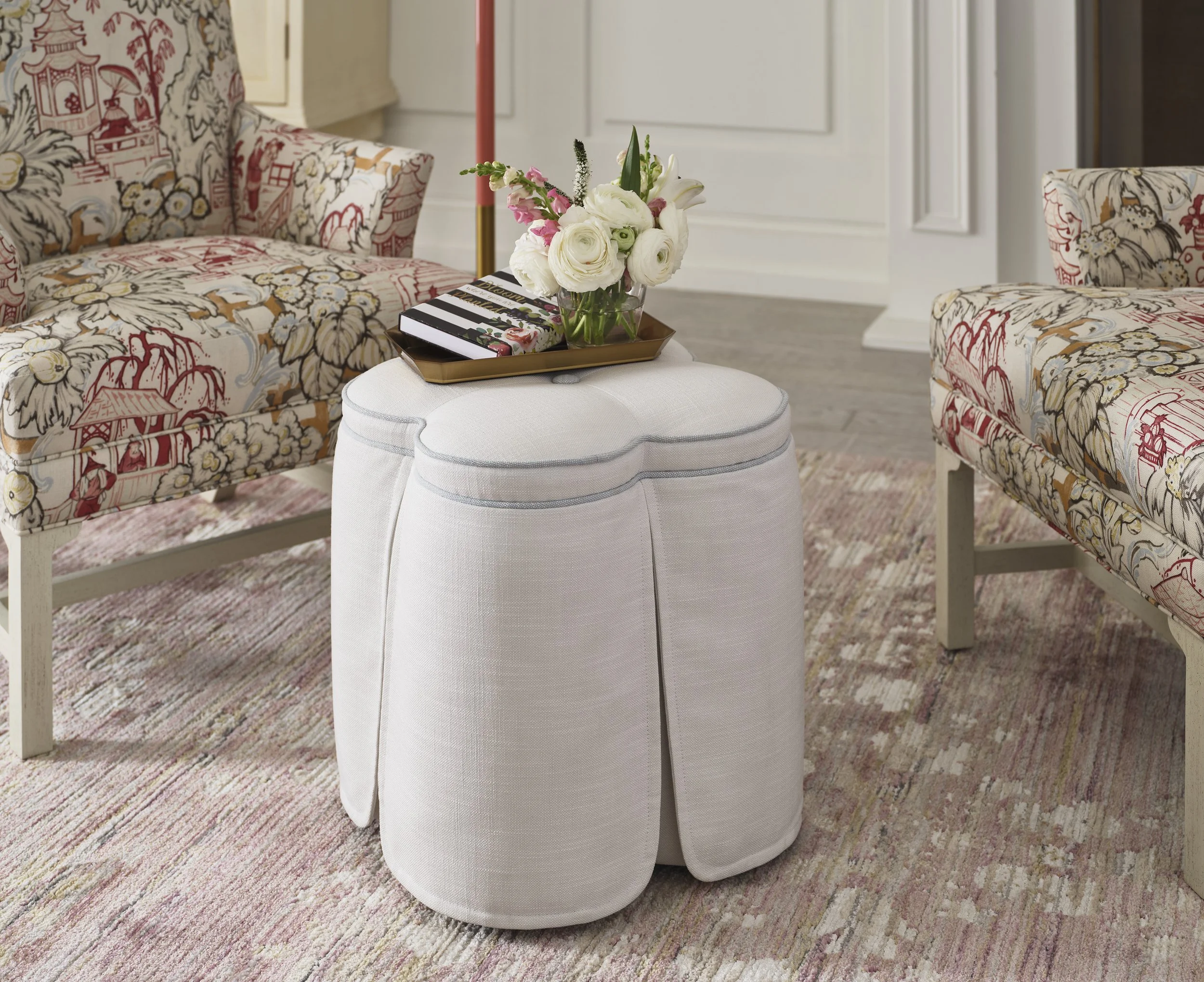A white upholstered ottoman with a vase of white and pink flowers, a tray, and a striped book on top, situated between two floral-patterned armchairs in a living room with a multicolored area rug.