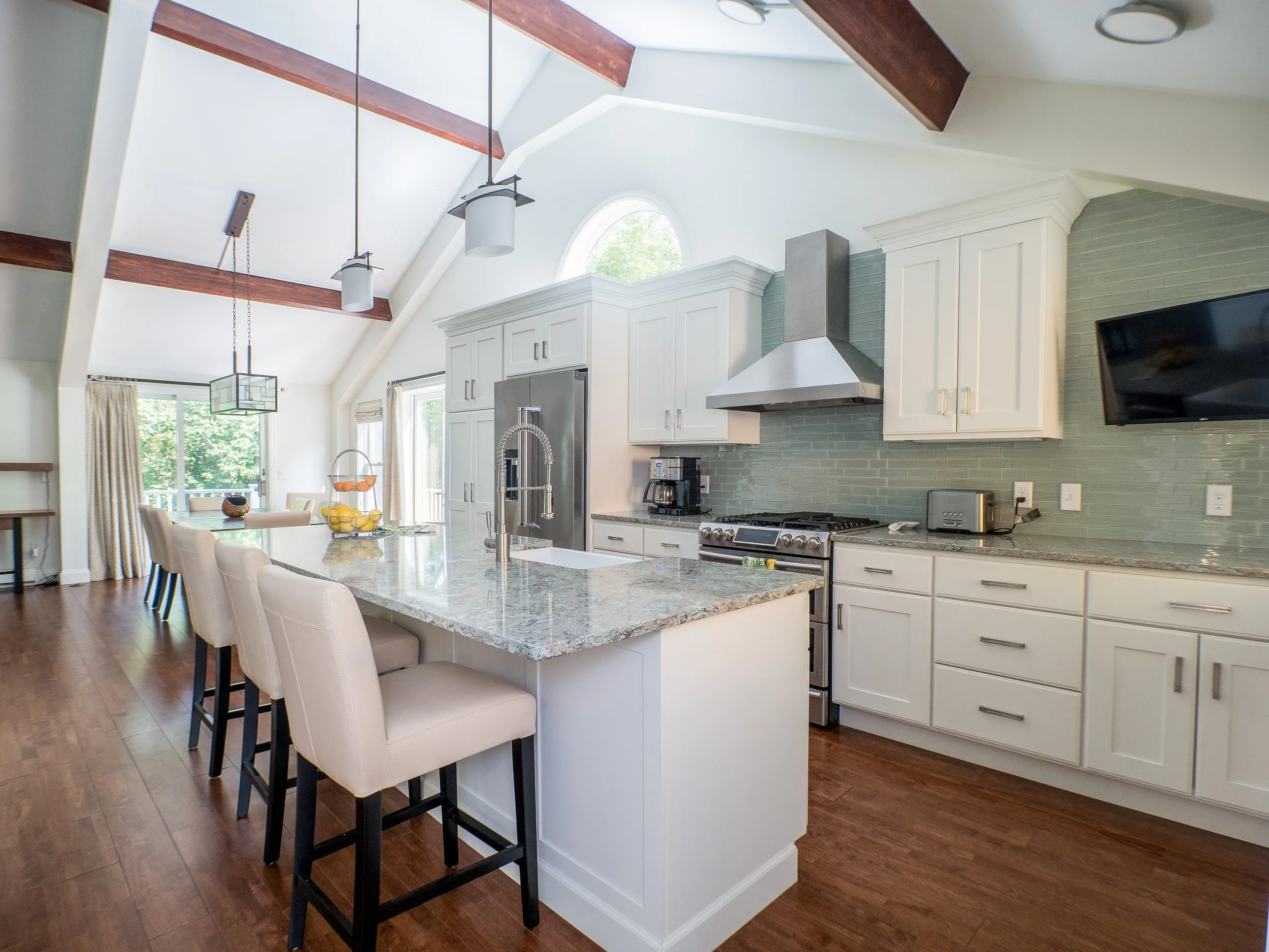 Bright kitchen with white cabinets, stainless steel appliances, a speckled granite island with four beige chairs, hardwood floors, and a high ceiling with exposed beams. Light green backsplash and a flat-screen TV on the wall.