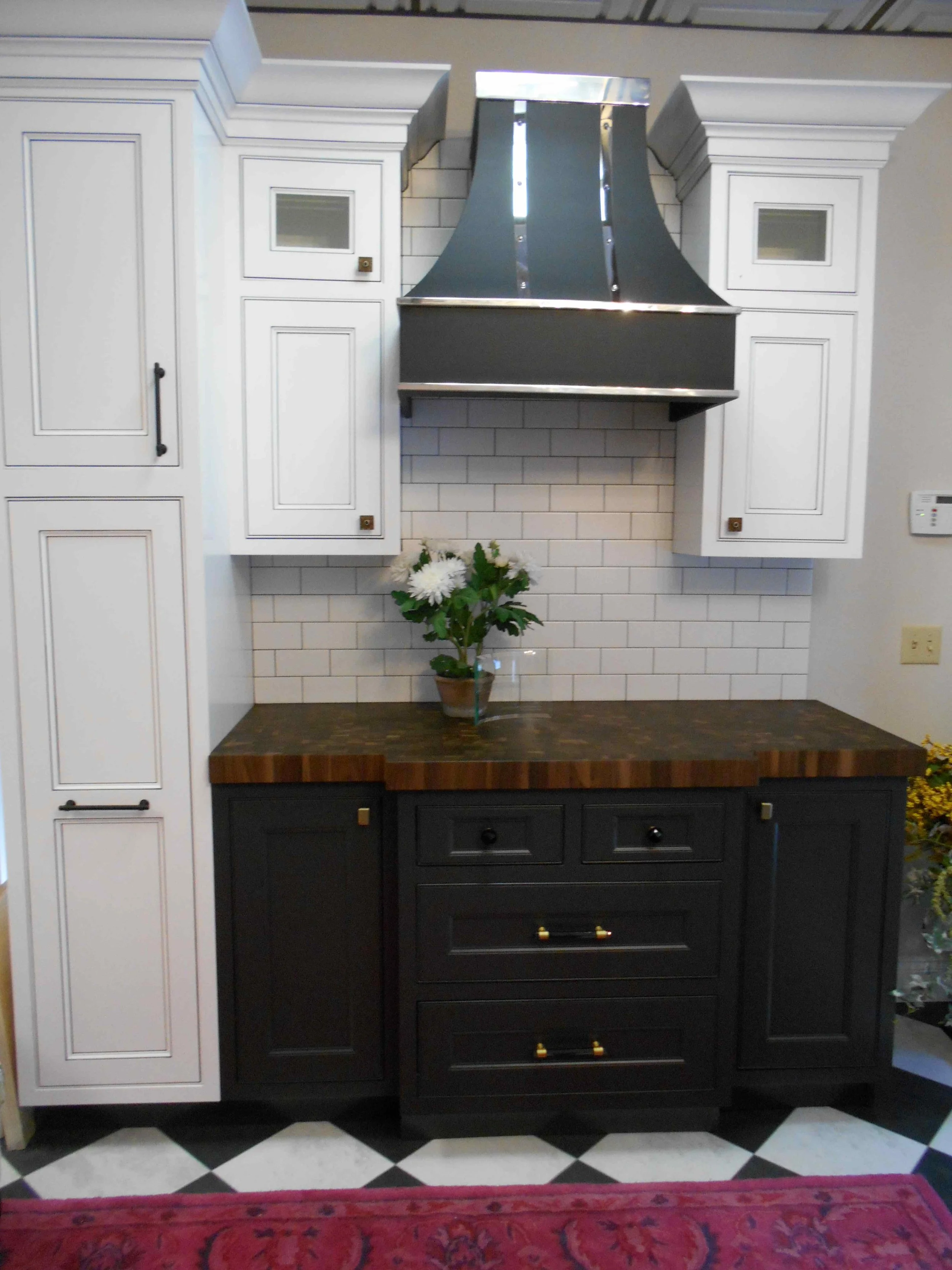 A kitchen with white upper cabinets, a black range hood, a dark wood countertop with a vase of white flowers, and black lower cabinets with gold handles. The backsplash is white subway tiles, and the floor has a black and white checkered pattern partially covered by a red rug.
