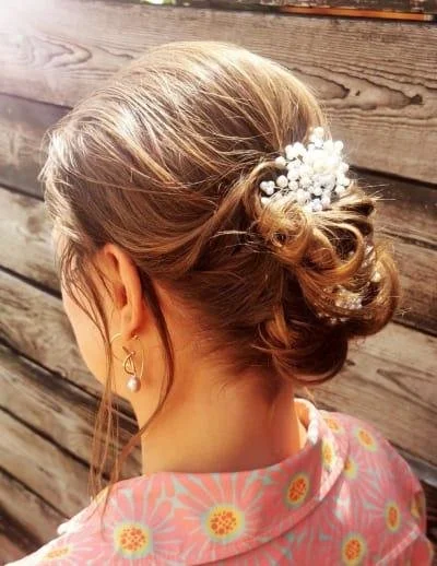 Woman with a styled bun hairstyle adorned with small white flowers, wearing gold earrings and a floral patterned shirt, standing in front of a wooden fence.