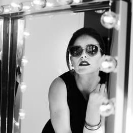 Black and white photo of a woman with sunglasses by a makeup mirror with lights.