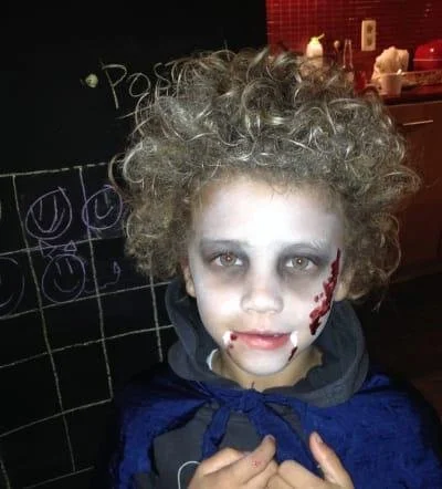 Child wearing zombie makeup with curly wig, in front of a chalkboard wall with doodles, inside a kitchen.