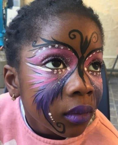Child with colorful butterfly face paint, featuring pink, purple, and black designs with white dots, looking upwards.
