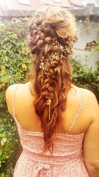 Woman with braided hairstyle adorned with floral hairpins, wearing a pink summer dress, stands in front of a garden wall.