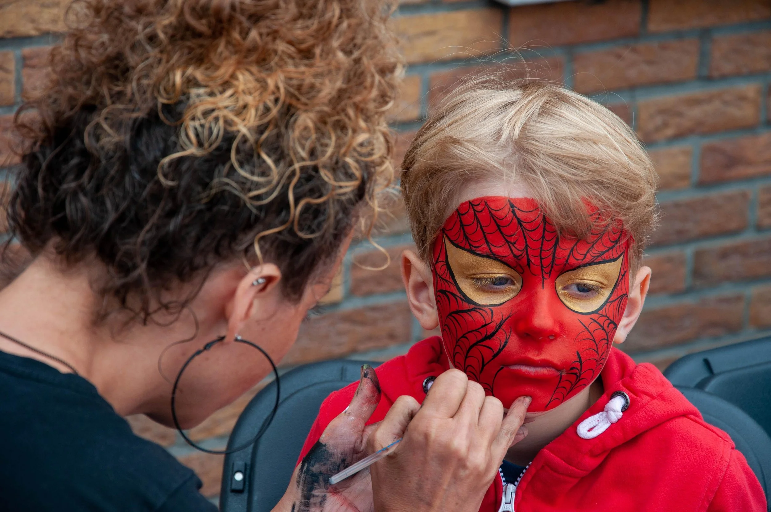 Young child with face painted like a superhero by artist in a red and black spider web design, wearing a red hoodie, face painting in progress.