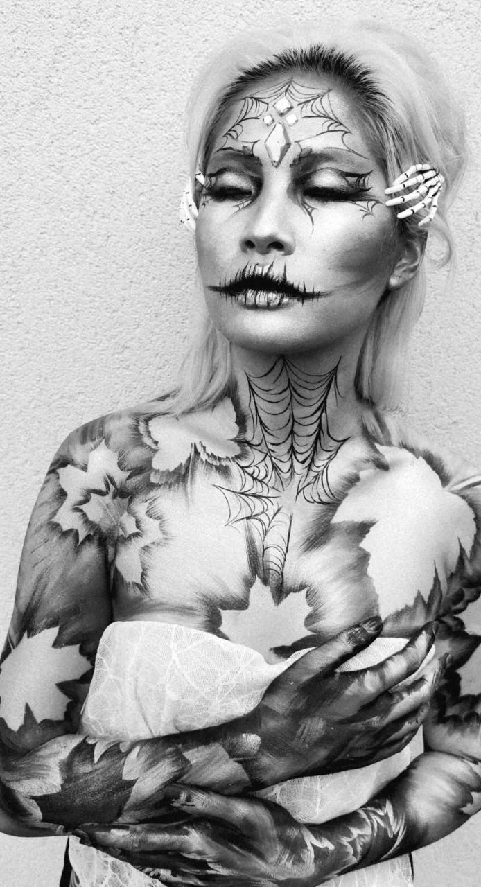 Person with intricate Halloween makeup and body art, depicting spider webs and skeleton patterns, in black and white.