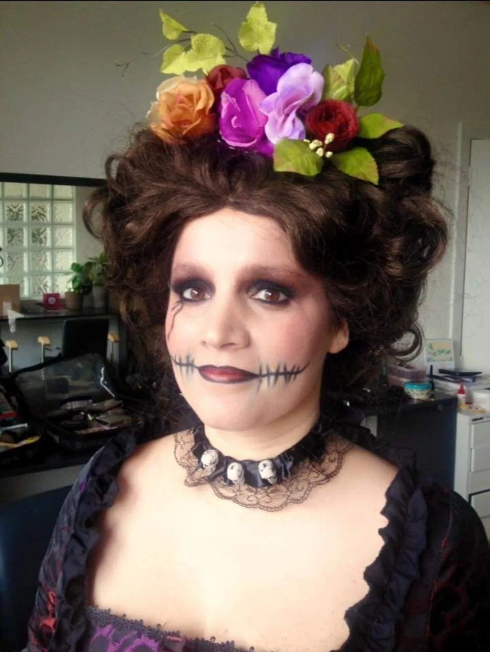 Person with colorful flower headpiece and face makeup resembling stitched lips, wearing a lace choker with skulls in a room with a table and plants.
