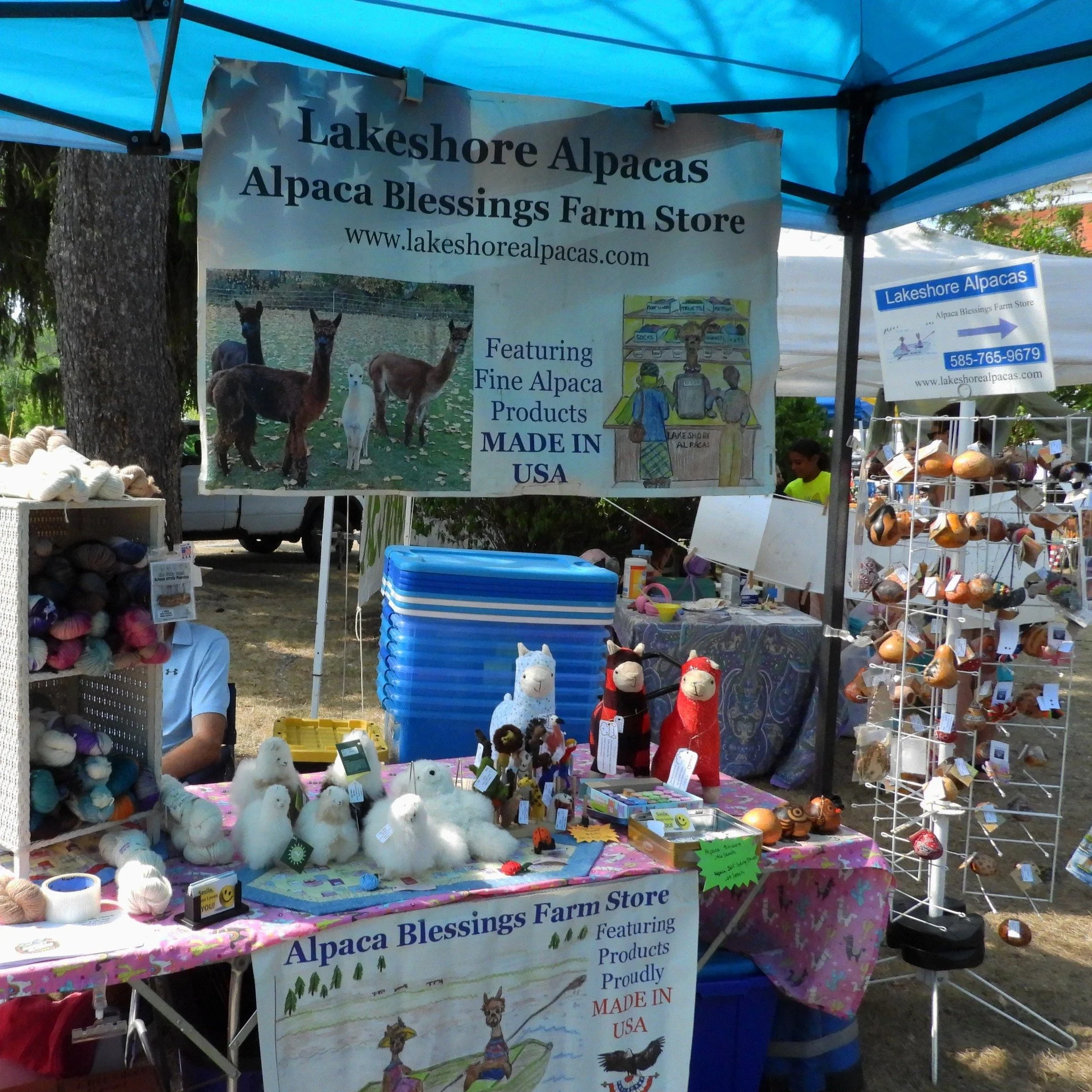 Tent at an outdoor craft fair displaying stuffed alpacas, yarn, and painted rocks, with signs promoting Lakeshore Alpacas and their products.