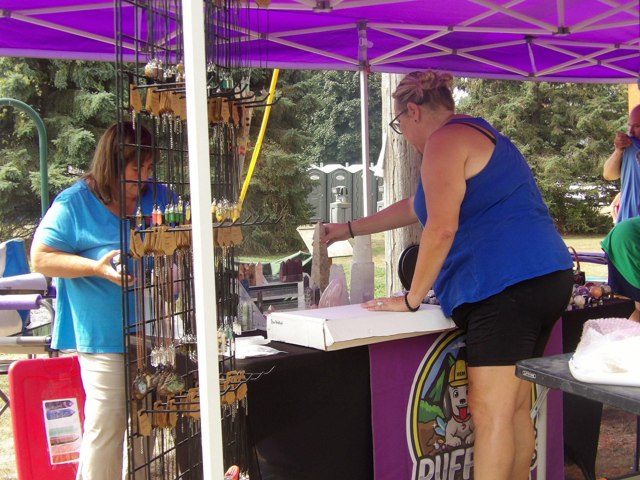Two women at an outdoor market stall with purple canopy, one browsing jewelry and the other arranging items, with outdoors trees and portable toilets in the background.