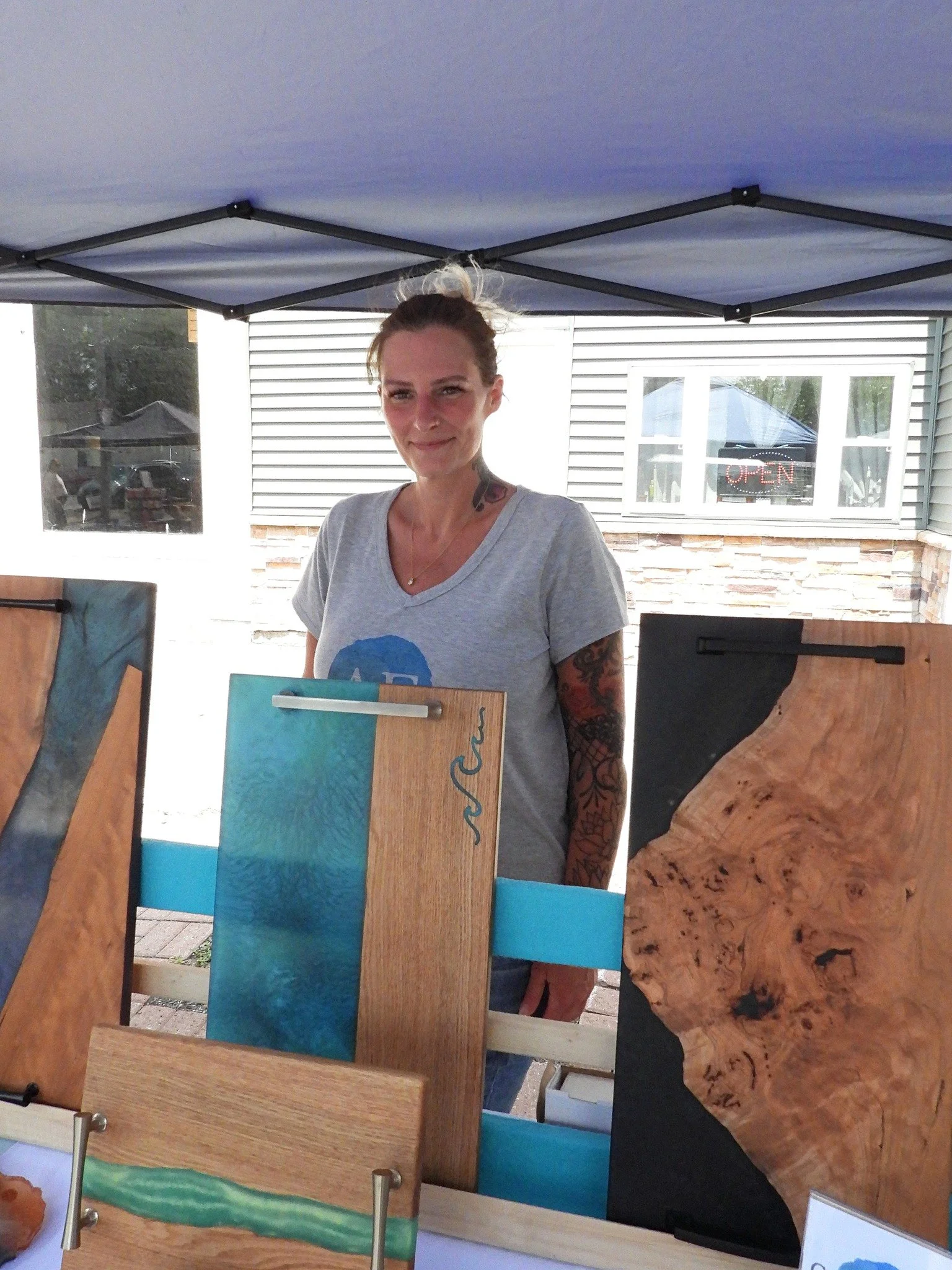 Woman with tattoos displaying wooden art pieces at an outdoor craft booth.