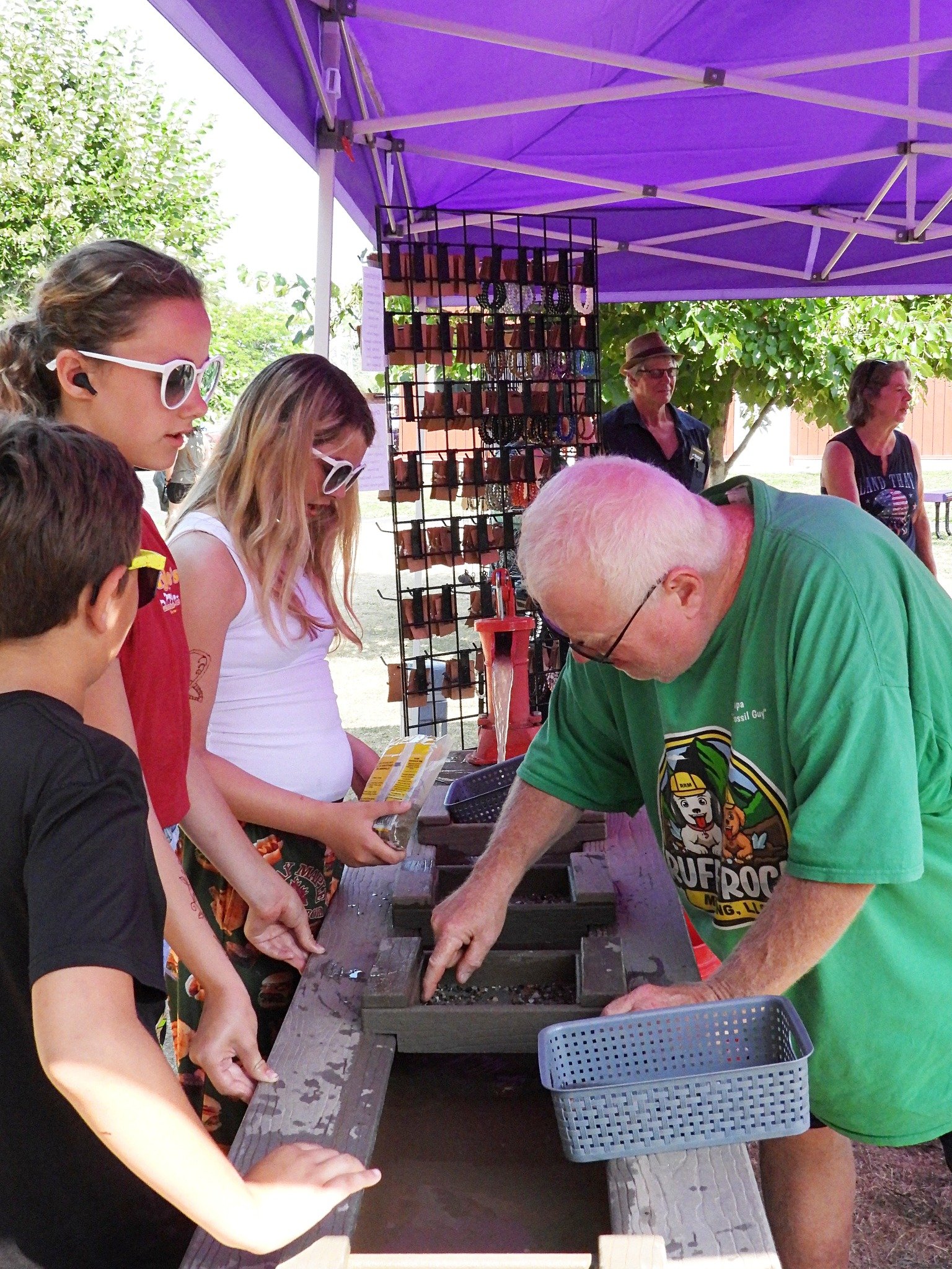 A man demonstrates a mineral or gem sorting activity at an outdoor event with several children observing. There are display racks with necklaces or jewelry and other items hanging in the background under a purple canopy.