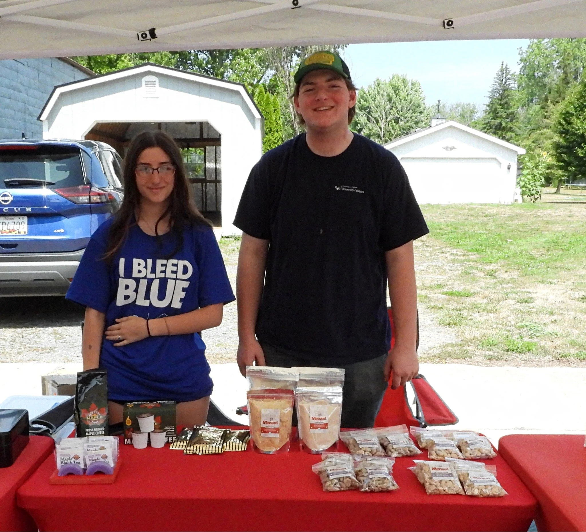 Two teenagers standing behind a red table under a white canopy, selling baked goods and snacks outdoors. The girl on the left wears a blue t-shirt that says 'I BLEED BLUE' and the boy on the right wears a black t-shirt and a green cap, smiling. There