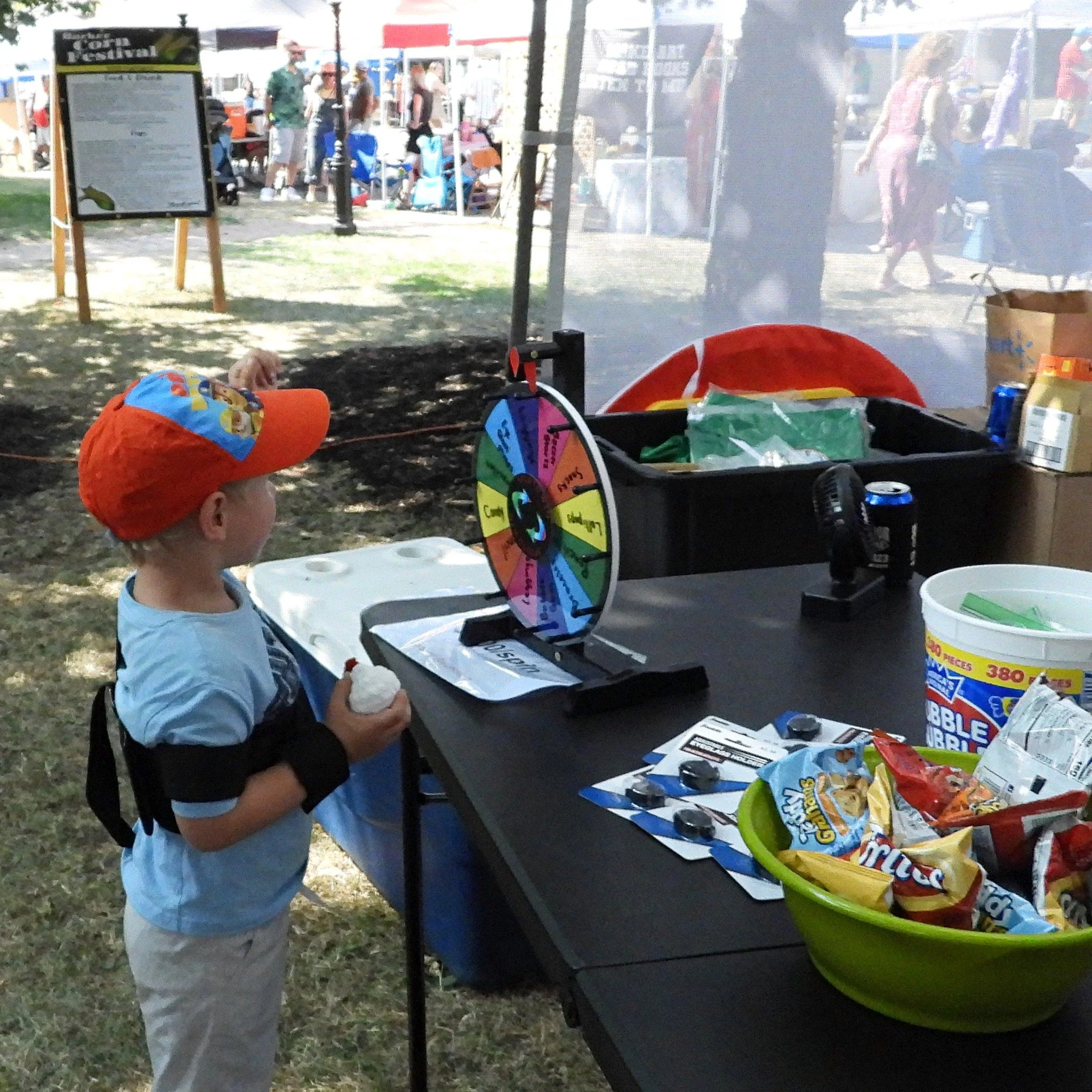 A young boy is standing at a carnival game booth with a colorful spinning wheel, holding a ball in his hand. The booth has snacks, drinks, and a prize bucket on the table. Other festival attendees are visible in the background.