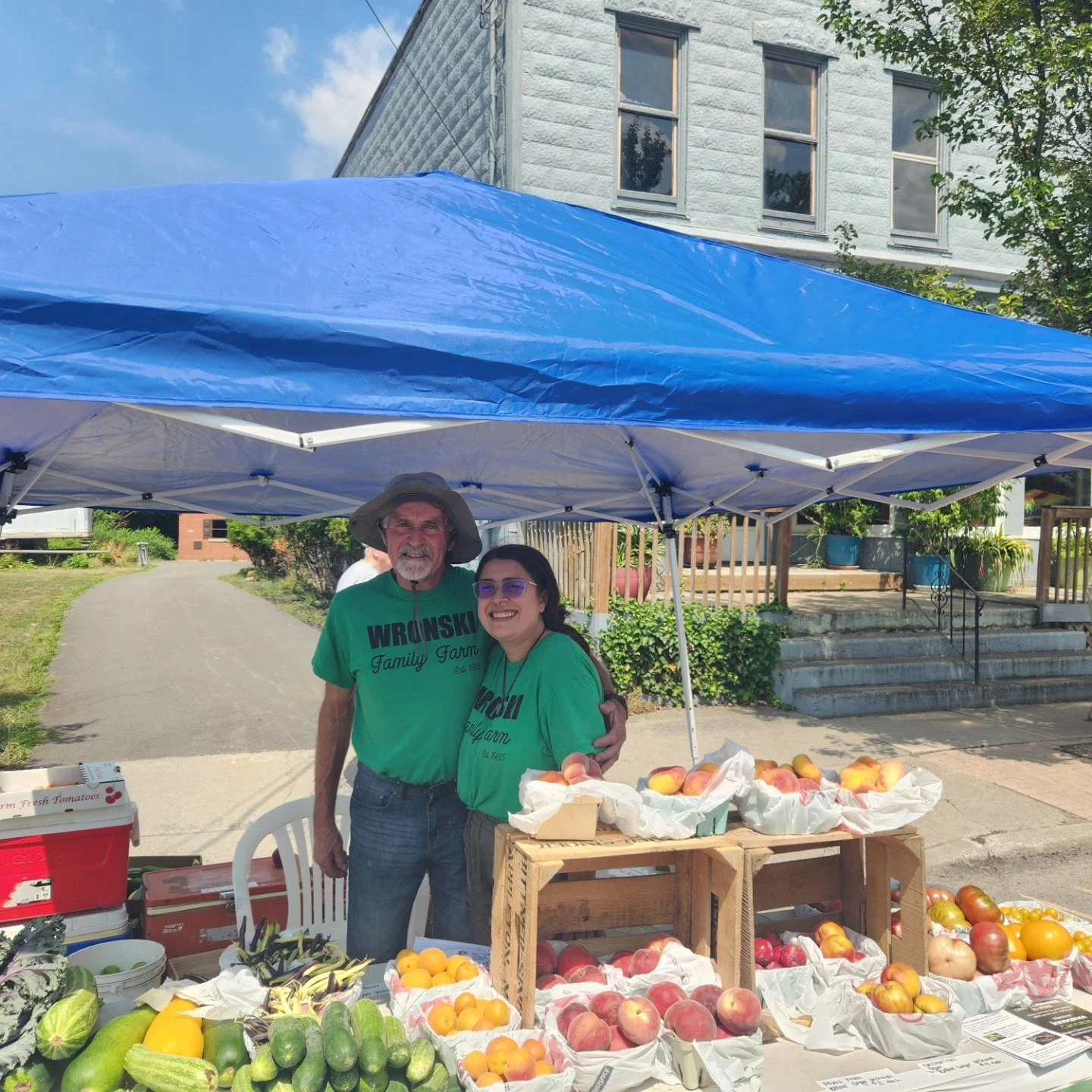 Two people standing under a blue canopy at a farmers market stall, selling various fresh fruits and vegetables. The man wears a wide-brimmed hat and a green T-shirt with the words "WRÅNSKI Family Farm," while the woman wears sunglasses and a matching