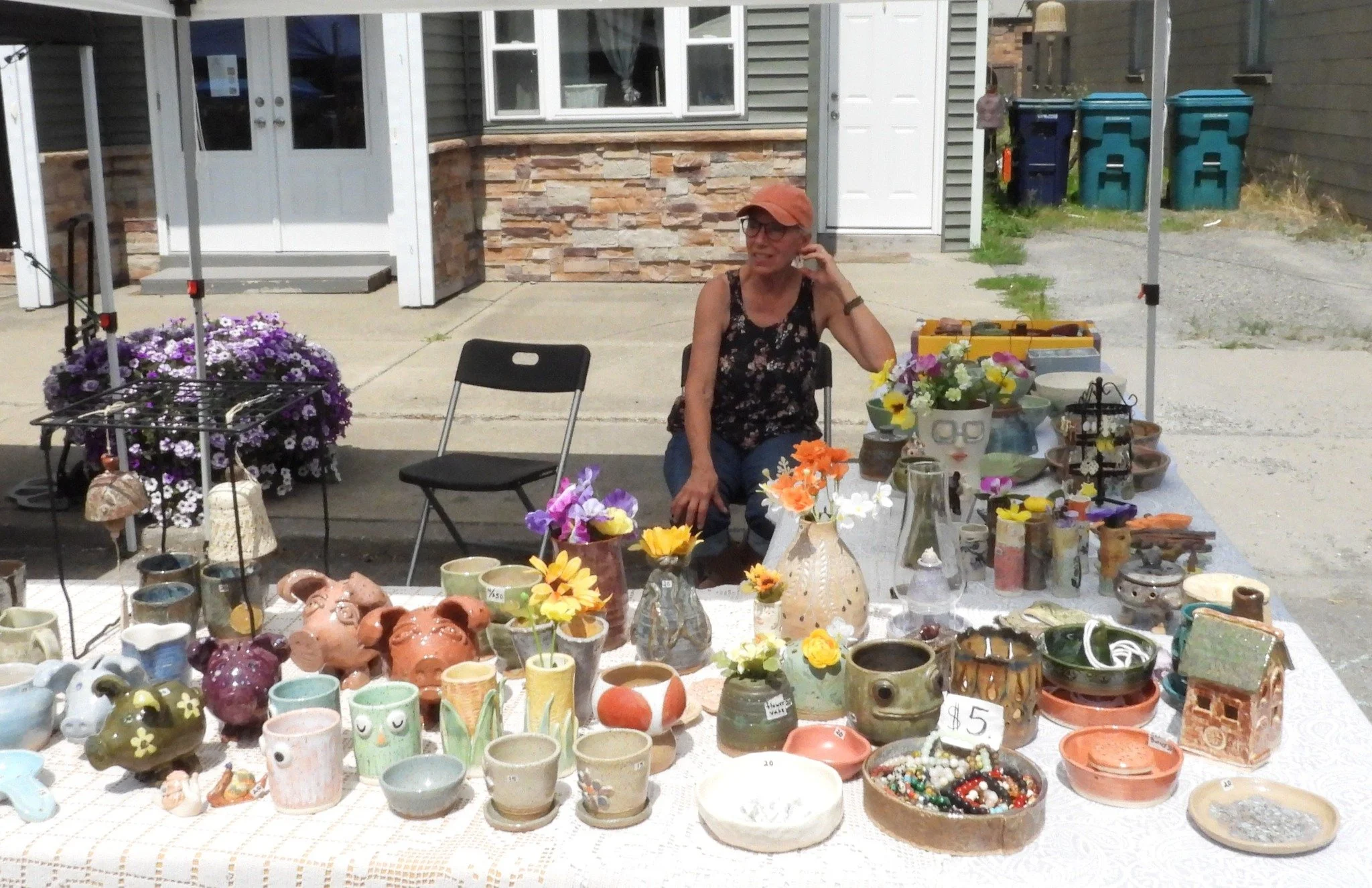 A woman sitting at a table with handmade pottery and flowers at an outdoor market. There are chairs, a house with a stone and siding facade, and various trash bins in the background.