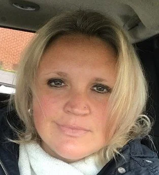 Close-up of a woman with blonde hair, wearing a white scarf, sitting inside a vehicle.