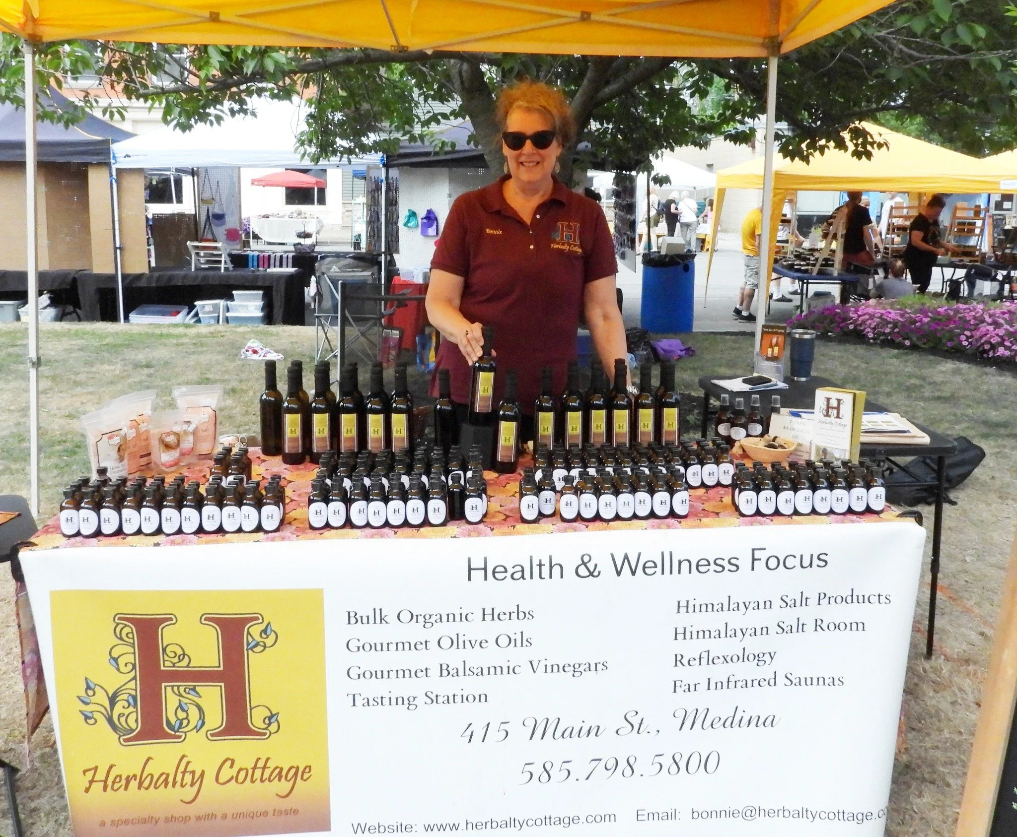 A woman standing behind a table at an outdoor market stall, selling health and wellness products. The table displays bottles and jars, with a sign listing various herbal and salt products. The woman is wearing sunglasses and a maroon polo shirt with 