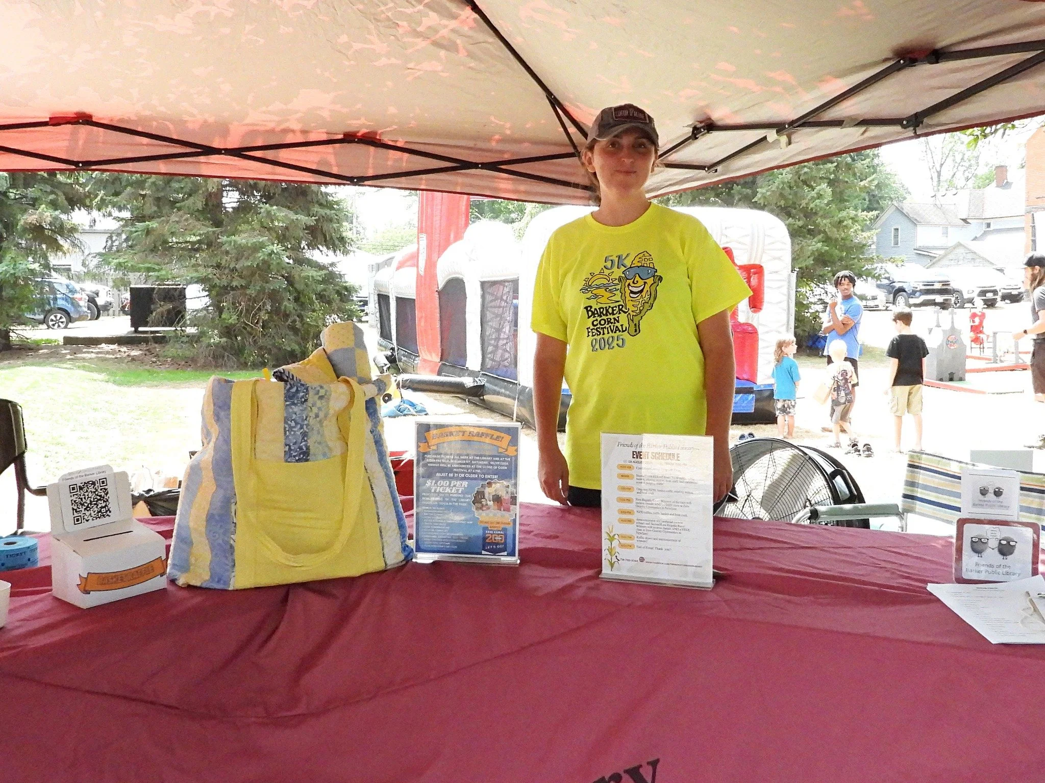 A young woman standing behind a table at an outdoor event under a canopy, with trees and other people in the background.