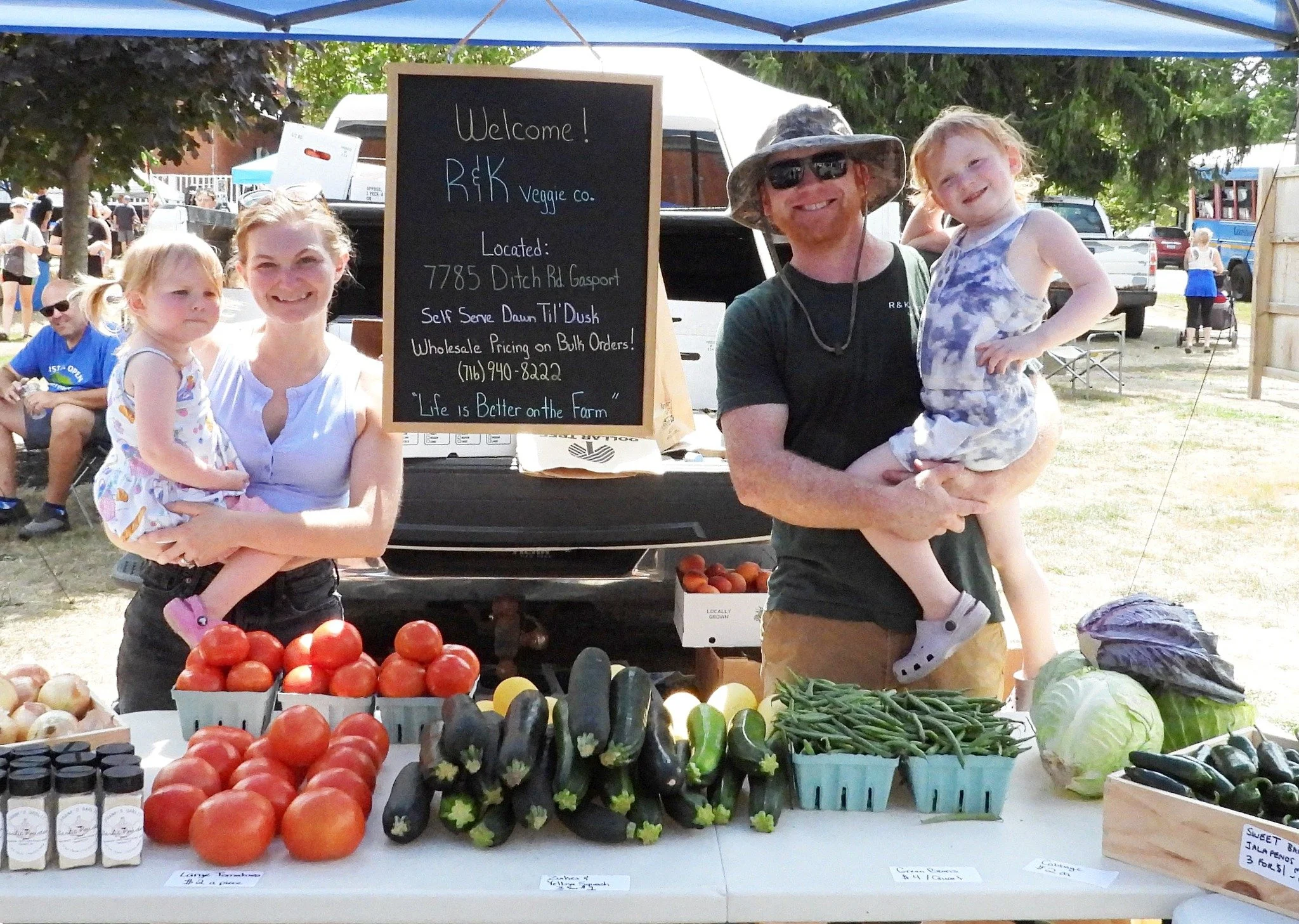 Family at a farmers market booth selling vegetables, including tomatoes, zucchini, cucumbers, and green beans, under a blue canopy with a sign indicating wholesale prices. The family members are smiling and posing for the photo.