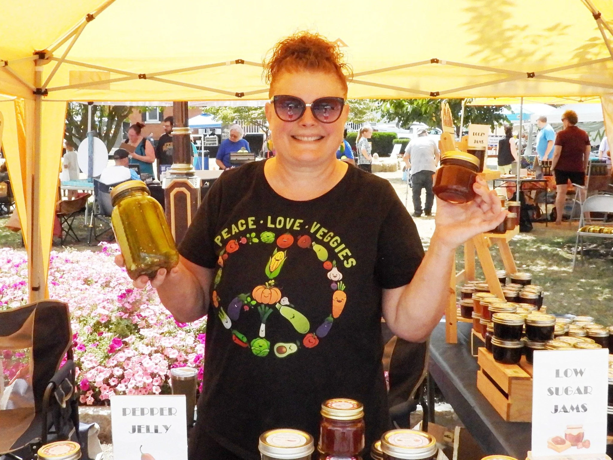 Woman at an outdoor market stall holding jars of pepper jelly and low sugar jams.