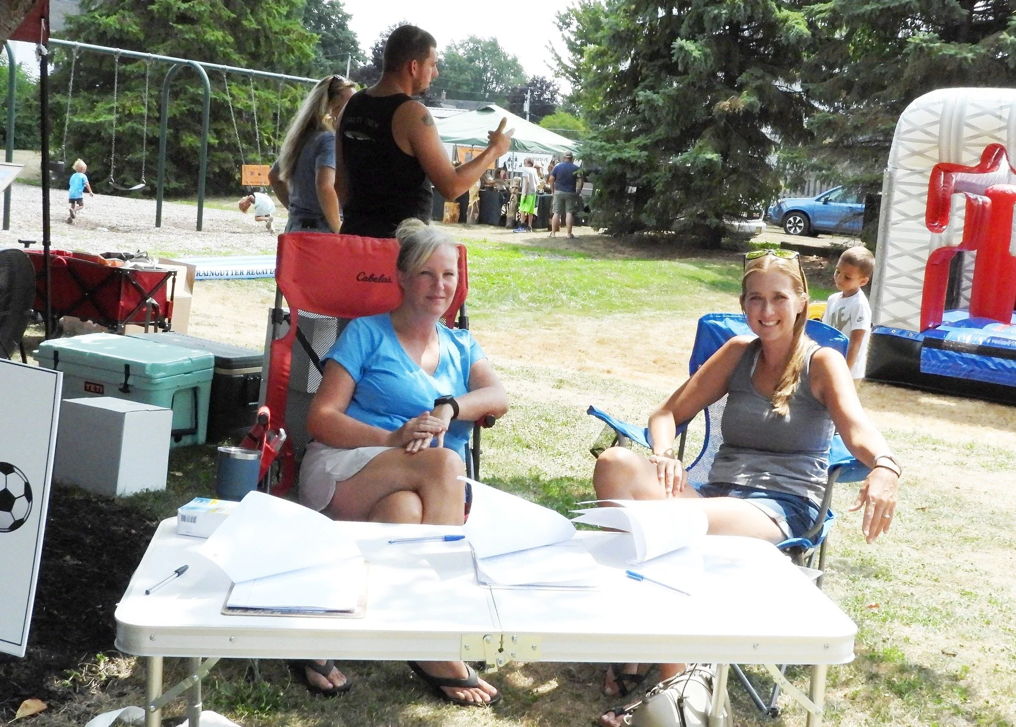 Two women sitting outdoors at a table with papers and pens, smiling at the camera during a community event or fair, with children playing and booths in the background.
