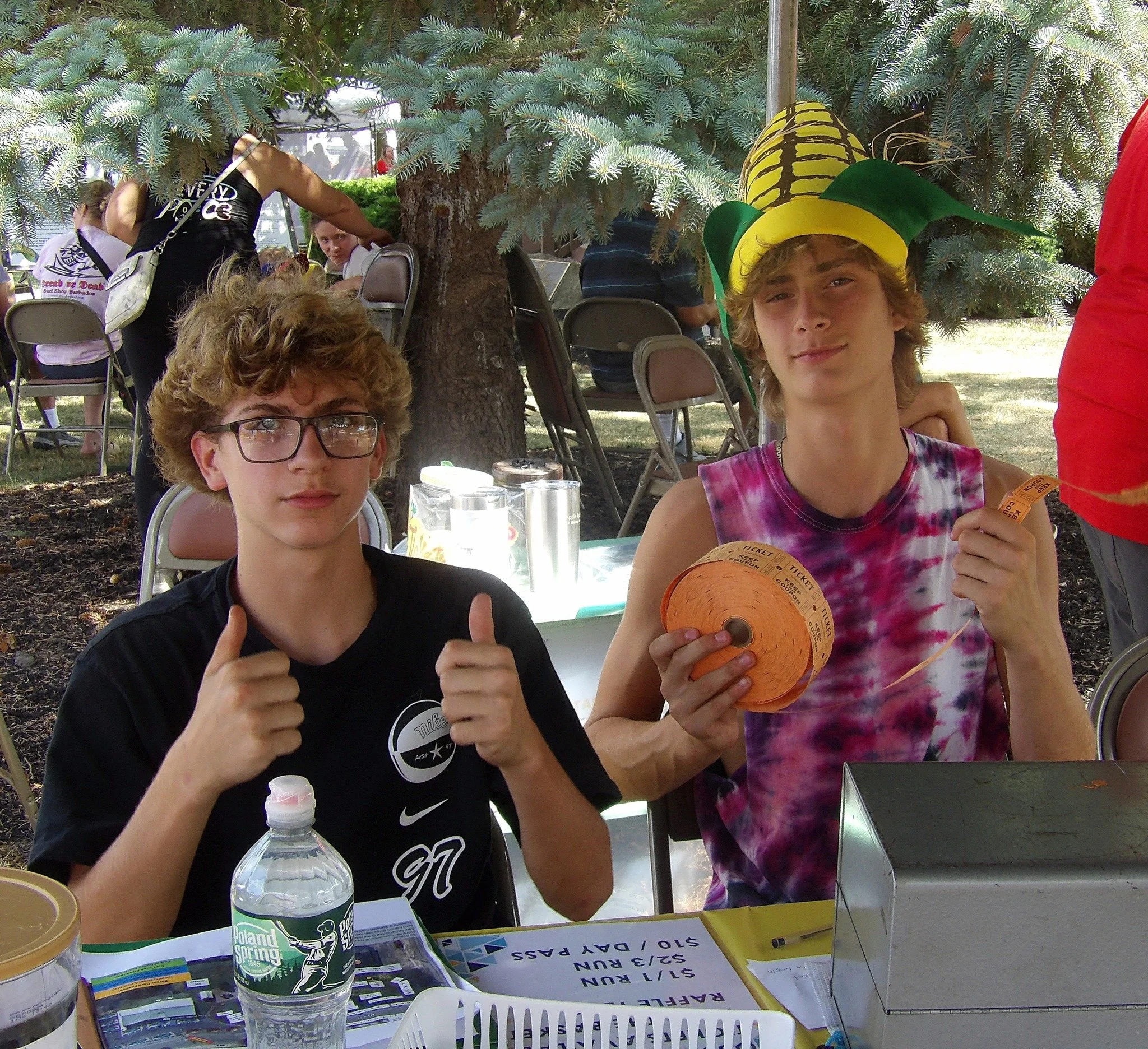 Two teenage boys sitting at a picnic table outdoors under trees at a fair or festival. One is giving a thumbs-up, wearing glasses and a black shirt. The other is wearing a colorful tie-dye shirt and a yellow and green hat, holding a lottery ticket an