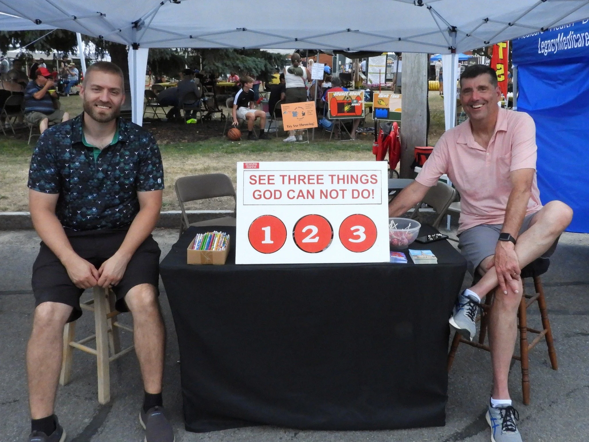 Two men sitting at a table outdoors at a community event under a white canopy. The table has a sign with the text 'See three things God cannot do!' followed by the numbers 1, 2, and 3 in red circles. One man is on the left wearing a dark, patterned s