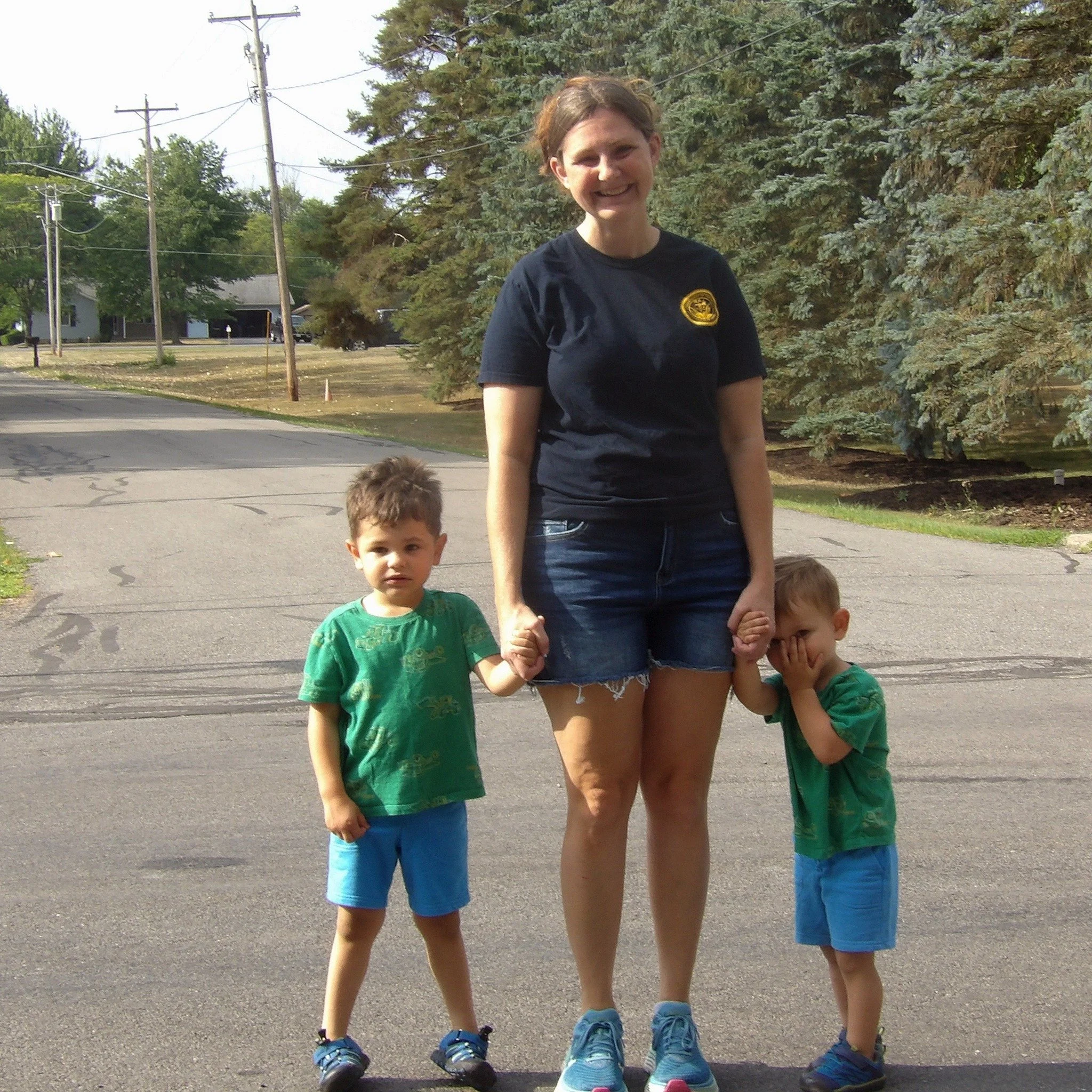 A woman with short brown hair wearing a black T-shirt and denim shorts is holding hands with two young boys on a quiet street with trees and houses in the background. The boy on the left has brown hair, is wearing a green T-shirt and blue shorts. The