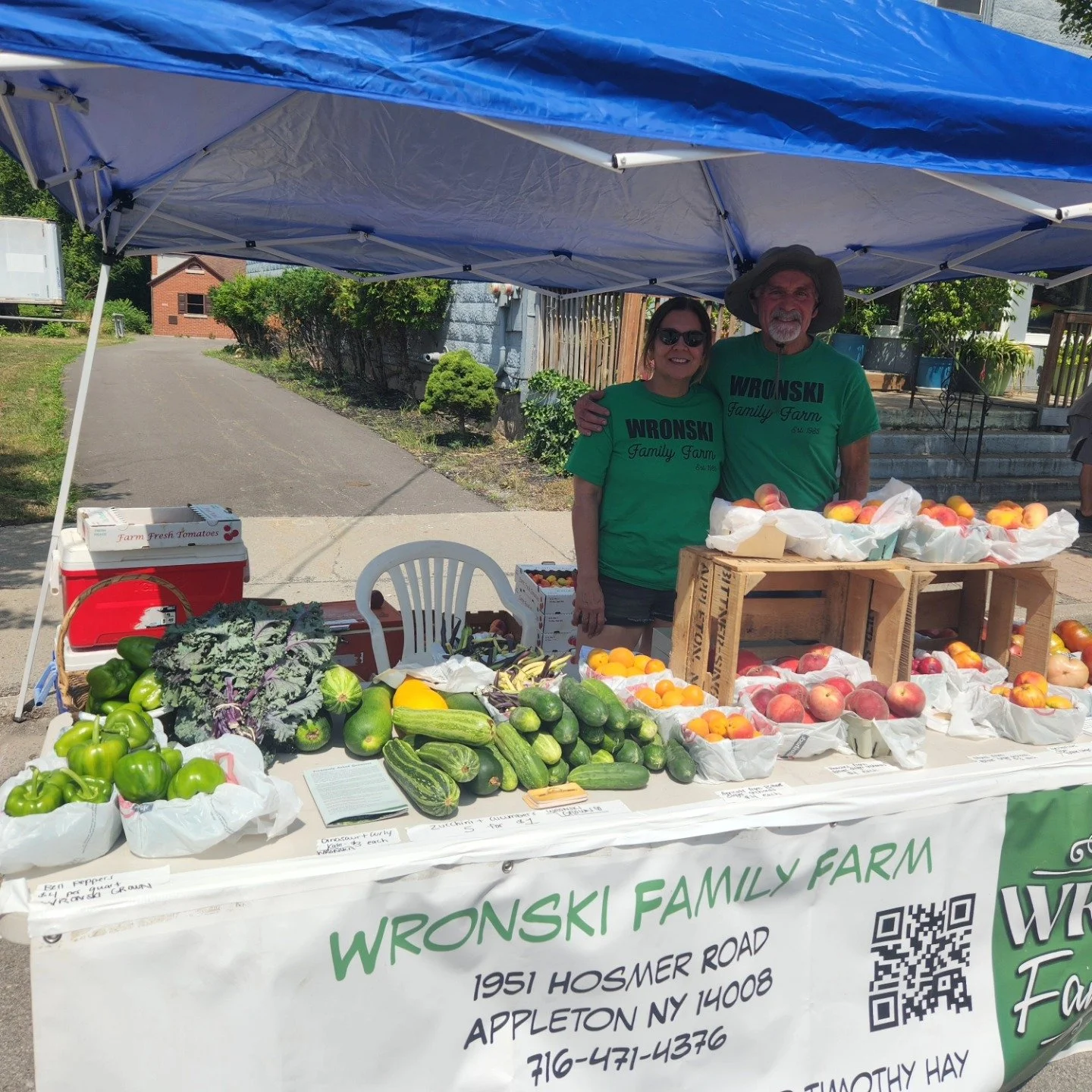 Two people standing behind a table of fresh vegetables and fruits at a farmers market stand under a blue canopy, with a sign that reads 'Wronksi Family Farm' and an address in Appleton, New York.